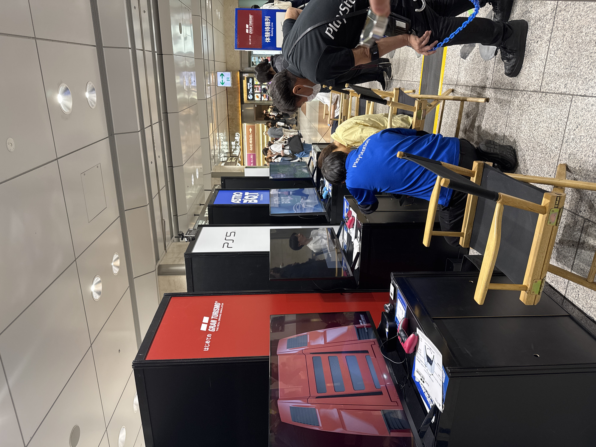 The image shows an indoor PlayStation event in Japan, possibly in Nakatsugawa. Several PlayStation 5 game stations are set up with titles like "Gran Turismo" and "Astro Bot." People are seated on director's chairs, engaging with the games. There are event staff members and signage, including "Gran Turismo" and "PlayStation" branding. A waiting line is visible in the background, along with general pedestrian traffic. The environment is a well-lit, tiled floor area, likely within a transportation hub, such as a train station or airport.