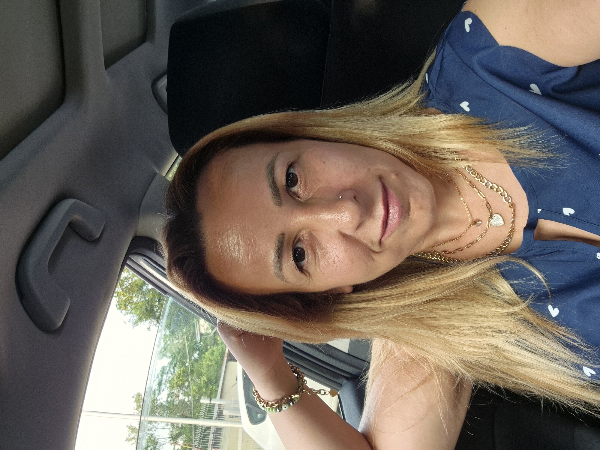 A woman with blonde hair and light skin, wearing a blue top with white hearts and multiple gold necklaces, takes a selfie in a car. She is leaning her head on her hand, with her arm resting on the car door. Her makeup includes lipstick and eyeliner. A bracelet with green beads and a gold chain adorns her wrist. The background shows the car interior, including the headrest and ceiling, as well as greenery and a fence visible through the car window.