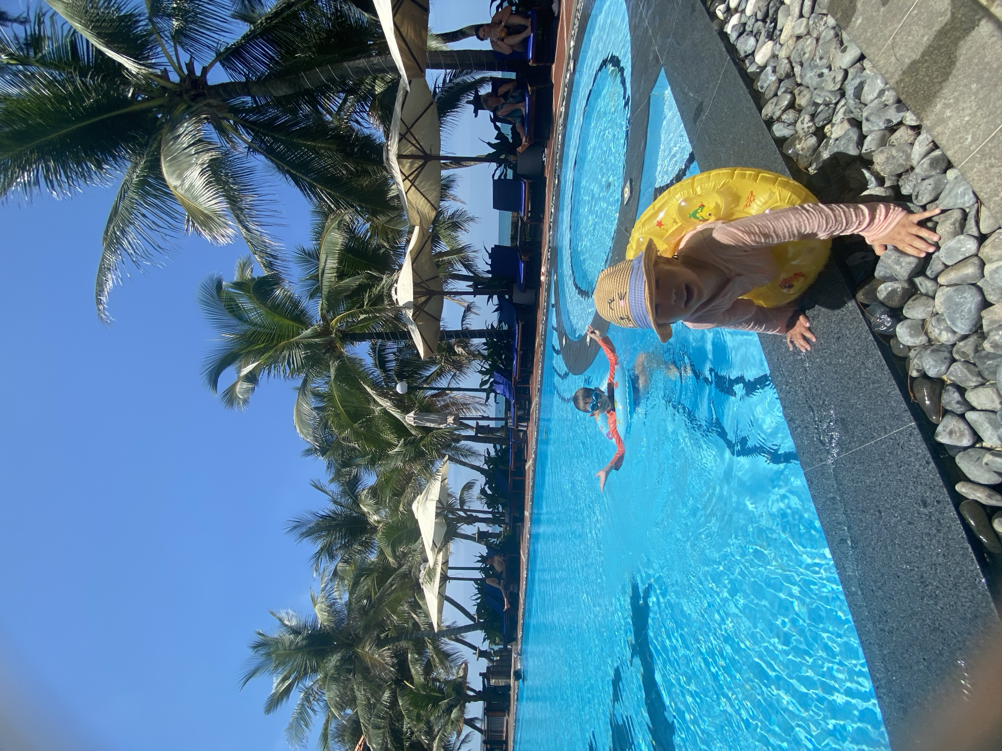 In this image, a clear blue swimming pool dominates the foreground. Two children are visible in the pool. One child, wearing an orange inflatable ring and goggles, is in the water. Another child, wearing a pink long-sleeved shirt, a straw hat, and a yellow inflatable ring, is at the edge of the pool, appearing to climb out. Palm trees are abundant around the pool, with lounge chairs and umbrellas visible in the background. The scene suggests a tropical resort setting in Phu Thuy Ward, Vietnam, under a bright blue sky.