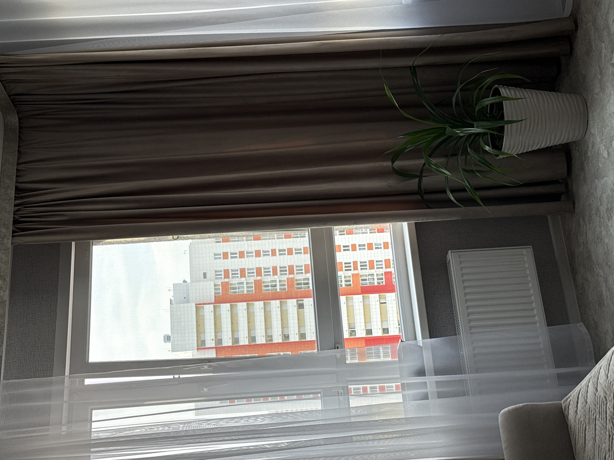 An interior view of a room shows a window with two curtains, one sheer white and one opaque brown, framing a view of an apartment building in Kazan, Russia. The window is fitted with a white radiator below it. A potted green plant stands to the right of the window, partially obscured by the brown curtain. A section of a sofa or armchair is visible in the lower left corner of the frame. The room appears to be a living space, possibly a bedroom, with textured wallpaper on the wall behind the radiator. The lighting suggests it is daytime, with natural light filtering through the sheer curtain.