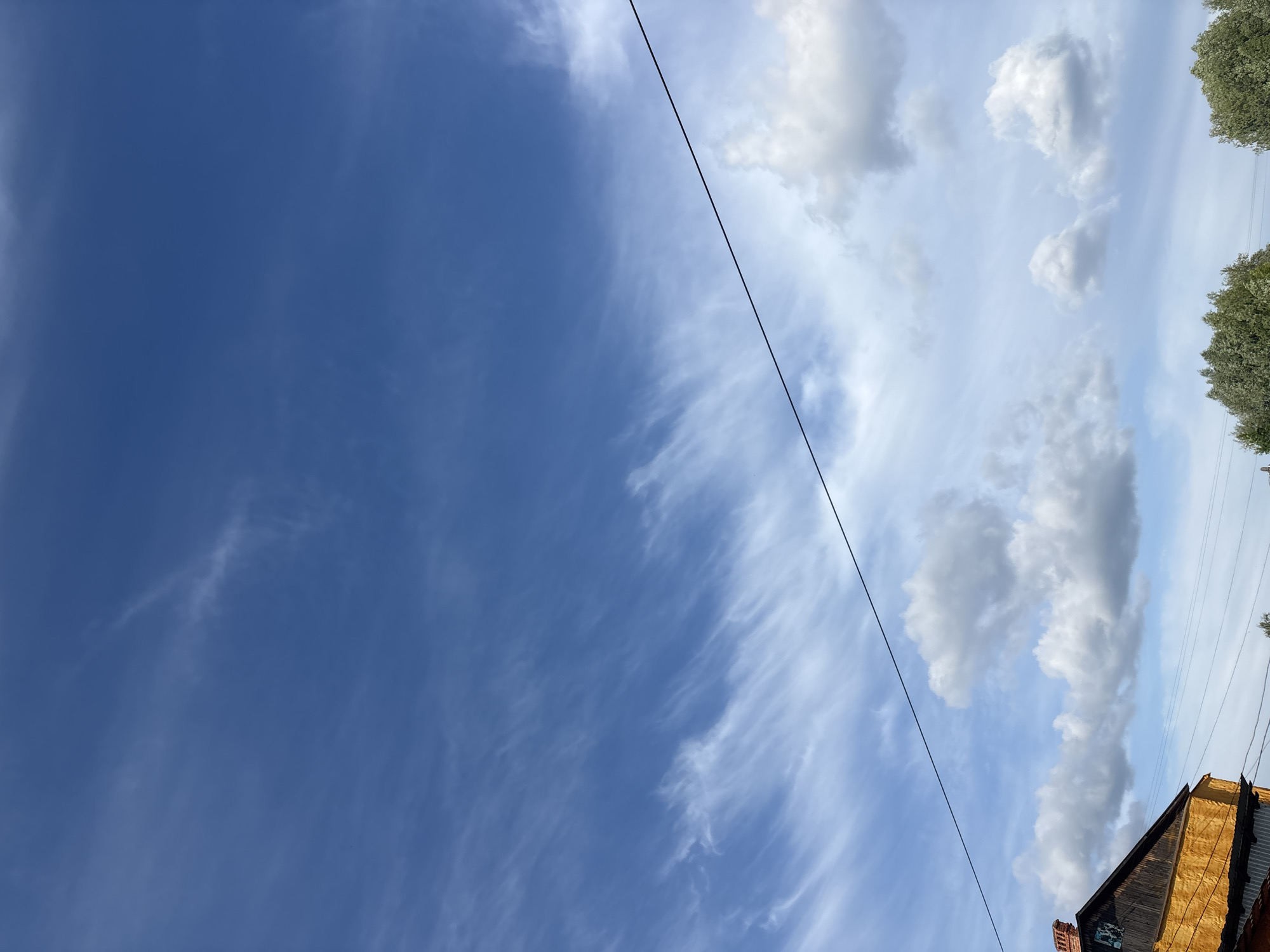An outdoor photograph, rotated 90 degrees counter-clockwise, primarily captures a bright daytime sky. The sky transitions from a deep, clear blue on the left to a lighter blue on the right, populated by various clouds. On the left side, the clouds are sparse and wispy, resembling cirrus formations. Towards the right, more substantial and fluffy cumulus clouds, in shades of white and light grey, dominate the sky.
A single, thin black power line stretches diagonally across the upper-mid section of the frame, extending from the top-left towards the bottom-right. In the upper-right corner, the vibrant green foliage of trees with light-colored leaves is partially visible. The lower-right corner of the image shows the upper part of a building, likely a house or barn, featuring a steeply pitched roof made of textured, possibly wooden, materials in warm tones of brown and orange. A small section of red brickwork, possibly a chimney, is also apparent.
The image depicts a clear, bright day with scattered clouds, indicating good weather conditions. There are no people visible, and no specific activity is taking place. No discernible text is present in the image. The setting appears to be rural or suburban, located in Фоки, Russia.
