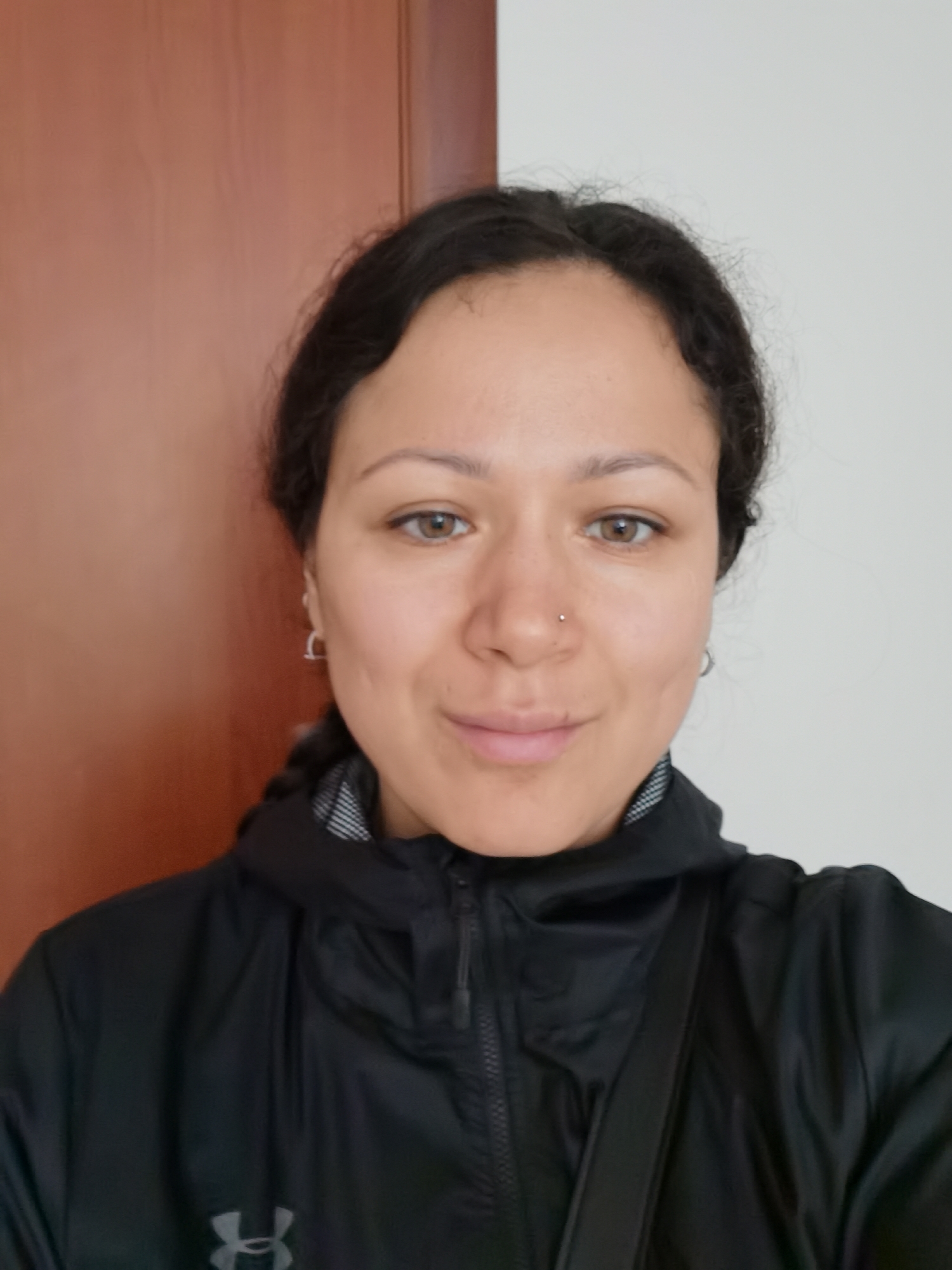 The image is a brightly lit, front-facing portrait of a woman, framed from the chest up. She appears to be in her late 20s or 30s, with fair skin, dark, wavy hair pulled back from her face, and warm brown eyes. A small silver stud piercing is visible in her right nostril, and she wears small silver hoop earrings in both ears. Her expression is neutral with a subtle, gentle smile.
She is dressed in a black hooded jacket, which has a gray and white patterned lining visible inside the hood. A black strap, possibly from a shoulder bag, is seen crossing over her right shoulder. On the left side of her jacket, the gray "Under Armour" logo is distinctly visible.
The background is an indoor setting, bifurcated into two sections. To the woman's left, a reddish-brown vertical surface, possibly a wooden door or paneling, is visible. To her right, the background is a plain, light gray or off-white wall. The lighting is even and soft, suggesting an indoor setting during daylight hours.