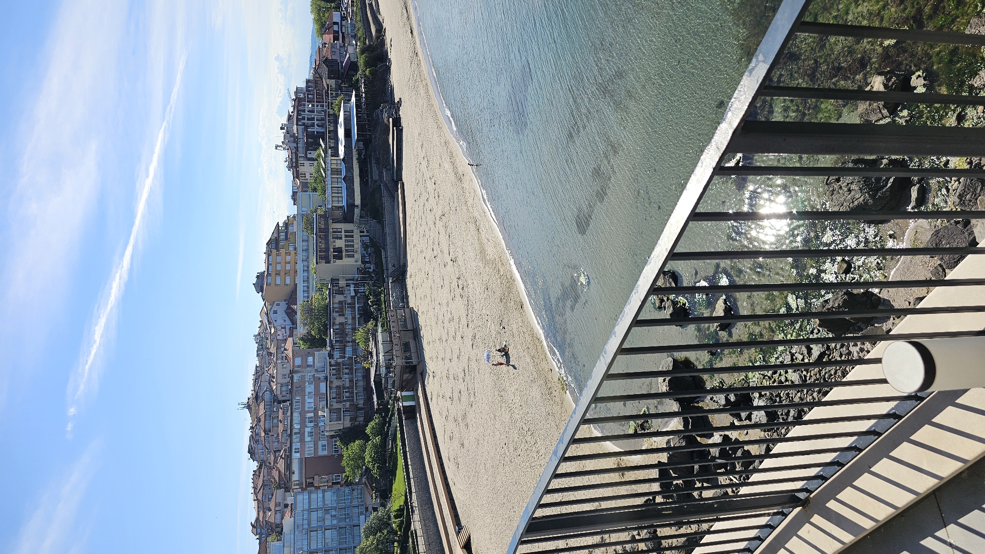 The image shows a view from a balcony overlooking a beach in Sozopol, Bulgaria. The beach has tan sand, and the water appears clear with a slight green tint. A few people are on the beach, and some are in the water.
On the left side of the beach, there is a row of buildings with a mix of residential and commercial properties. Some vegetation, likely trees and bushes, grows alongside the buildings. The sky is blue with scattered clouds, and the overall weather seems clear and sunny.
The balcony railing is visible in the foreground of the image. The railing is made of horizontal metal bars. Below the railing, rocks are visible in the water.