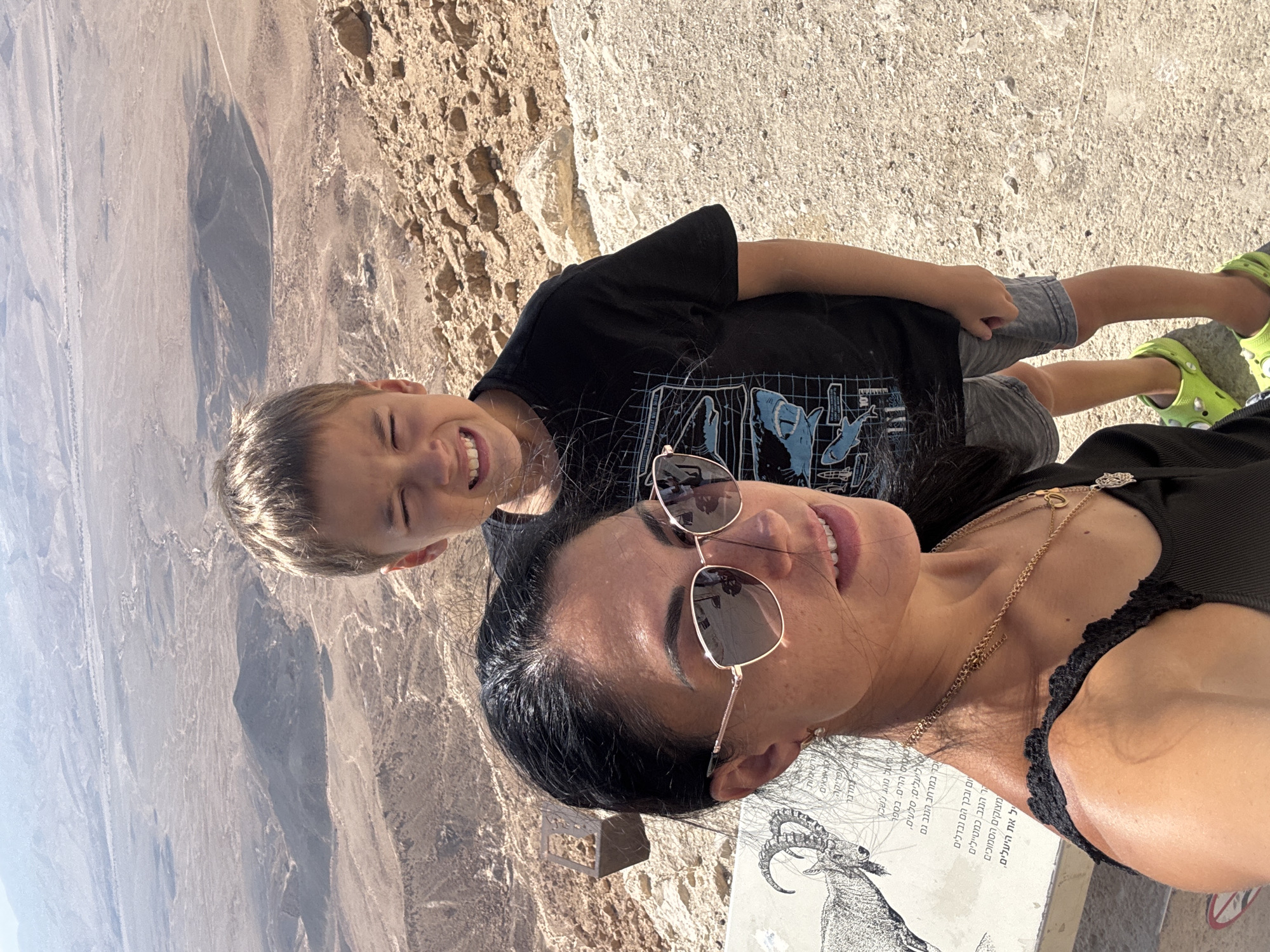 In Mitzpe Ramon, Israel, a selfie captures a woman and a young boy standing on a rocky, desert landscape overlooking a vast, arid valley. The woman, in the foreground, wears a black tank top, sunglasses, and a gold necklace. She has dark hair and is looking towards the camera with a slight smile. The boy, positioned behind and to her right, is wearing a black t-shirt with a blue graphic of sharks and rockets, grey shorts, and bright green Crocs. He is squinting and smiling widely at the camera. In the background, the expansive desert unfolds with hills, valleys, and dry riverbeds under a hazy sky. A sign with an illustration of an ibex and Hebrew text is partially visible to the left of the woman. The overall impression is a tourist snapshot in a natural, desert environment.