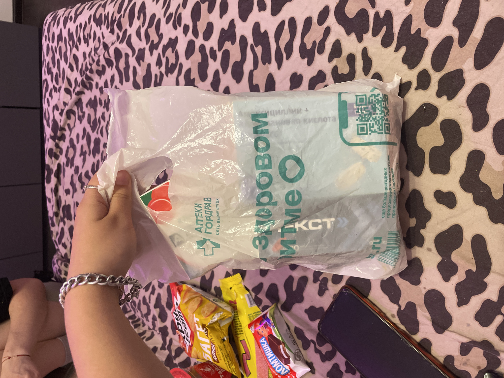 A close-up, high-angle, indoor shot shows a person's hand holding a white plastic bag with the green text "Аптеки Горздрав" and "здоровом мине" visible. Next to the bag are two snack bags, one yellow and one red with white text. A smartphone with a black screen and orange casing lies on a bed with a leopard print blanket. The person's left arm, wearing a silver chain bracelet, is visible in the upper left corner. The background is a bed with a leopard print blanket. The context of Irinovka, Russia is not discernible from the image.