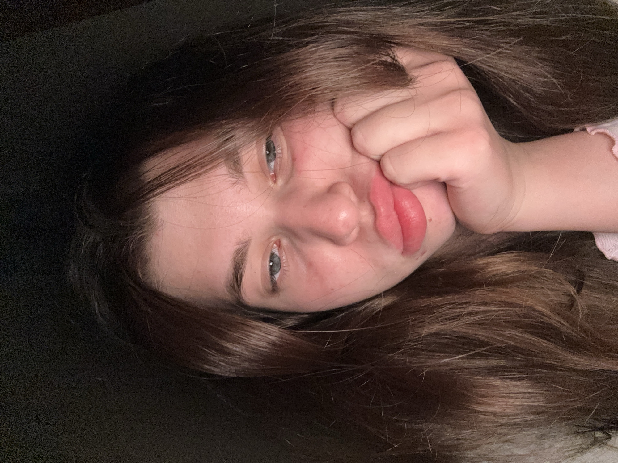 A young woman with brown hair and blue eyes is lying down, her face tilted to the side. Her hand is pressed against her cheek, distorting her lips into a pout. The background is dark, with a portion of black surface visible. A small part of what appears to be light-colored clothing is visible on the right side of the image. There is no information to suggest this image was taken in Мещёры, Russia.