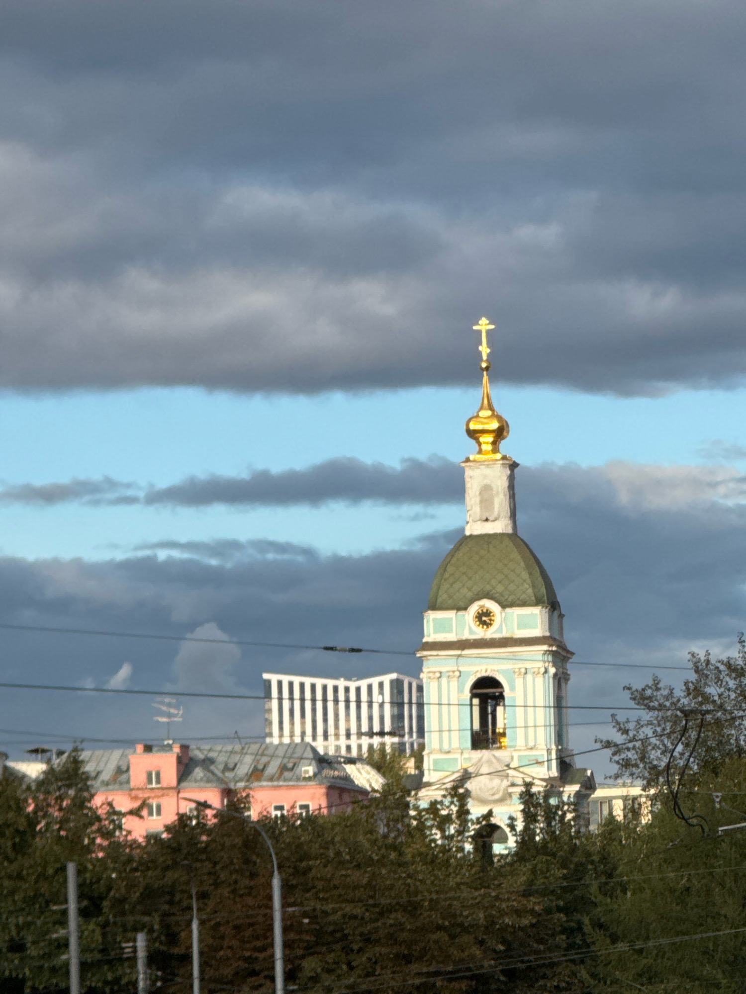 The image shows a view of the Church of the Resurrection of Christ at the Rogozhskaya Sloboda in Moscow, Russia. The church's bell tower, topped with a golden dome and cross, is prominent against a sky filled with dramatic, dark clouds interspersed with patches of blue. Below the bell tower, the main body of the church features a green, pitched roof and decorative light blue and white architectural elements, including a clock face. In the middle ground, a pink building with a grey, multi-pitched roof is visible, partially obscured by trees. Further back, a modern white building with vertical stripes stands out. The foreground is dominated by dense green trees and utility wires, suggesting an urban environment. The lighting indicates it is likely late afternoon or early evening, with the sun casting a warm glow on the church's golden dome.