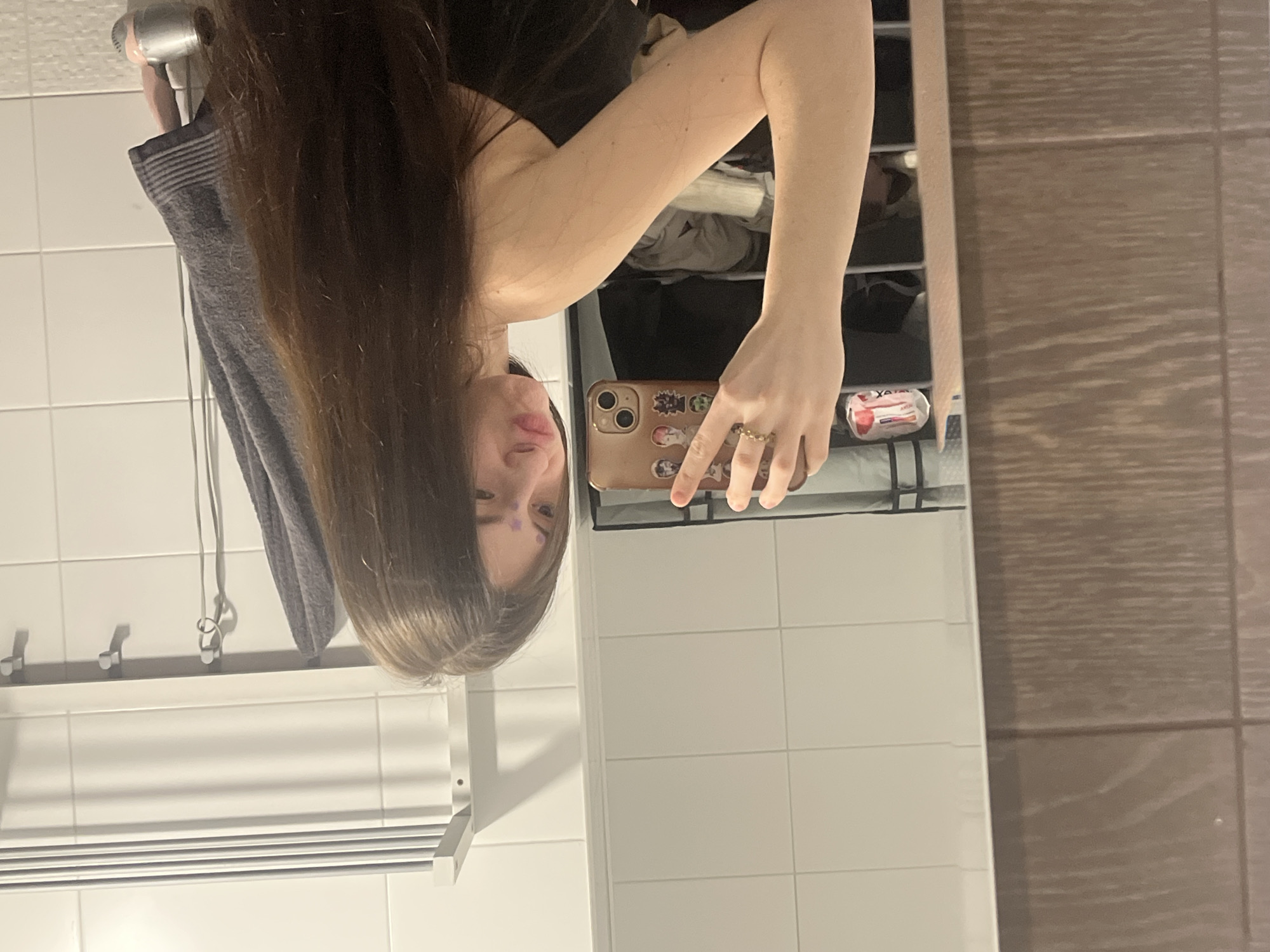 This media file captures a mirror selfie taken indoors, likely in a bathroom or dressing area, though the image is currently rotated 90 degrees clockwise.
The main subject is a person with long, dark brown hair, seen in reflection. Her face is partially visible, featuring a playful expression with puckered lips. Small purple marks, possibly makeup or decorative stickers, are visible on her forehead/eyebrow area. She appears to be wearing a dark top, such as a camisole or tank top.
She holds a rose gold or copper-colored iPhone, which has several colorful stickers on its back, depicting what appear to be animated characters. A delicate, golden, wavy-patterned ring adorns one of her fingers as she grips the phone.
The reflected background reveals a setting with both white and brown tiled walls. To the left of the person (when the image is correctly oriented), a white tiled wall features a white shelf with hooks, holding a grey towel and a silver chain or necklace. Below this shelf, a white slatted structure, possibly a towel warmer or radiator, is visible. Behind the person's head and upper body, a storage unit with multiple cubbies holds various folded items, likely clothing or linens. To the right, a brown tiled wall with a textured pattern adds contrast. A small, white and red package, with the word "Always" partially legible, is seen in the lower right corner of the reflected scene.
The activity is a casual mirror selfie. The time of day is not evident, as the scene is illuminated by indoor lighting. The overall mood is personal and candid.
