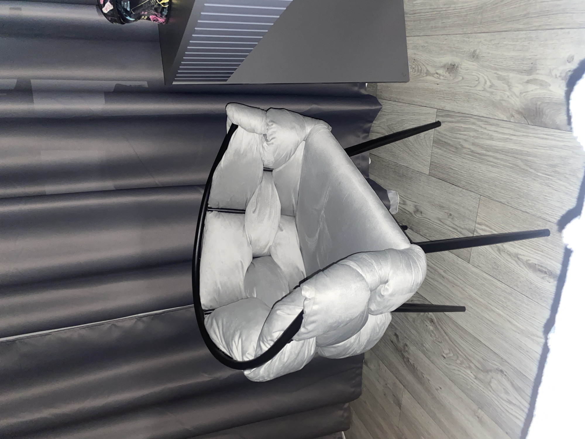 This is a medium shot, eye-level, indoor photograph of a light gray, tufted armchair with a black metal frame, situated on a light wood-grain laminate floor. The chair has a plush, rounded seat and backrest, with the fabric appearing to be gathered and knotted, creating a soft, pillowy effect. The chair has four thin, black metal legs.
Behind the chair, dark gray curtains are visible, drawn closed. To the right of the chair, a section of a dark gray furniture piece with vertical white lines is present. A small portion of a colorful, patterned object, possibly a hat or bag, is perched on top of this furniture.
The lighting is dim, casting some shadows, particularly a large shadow from the chair onto the floor in the foreground. The scene appears to be a room, likely a bedroom or living space, given the furniture and curtains. There are no people visible in the image. The location is identified as Krasnoyarsk, Russia.