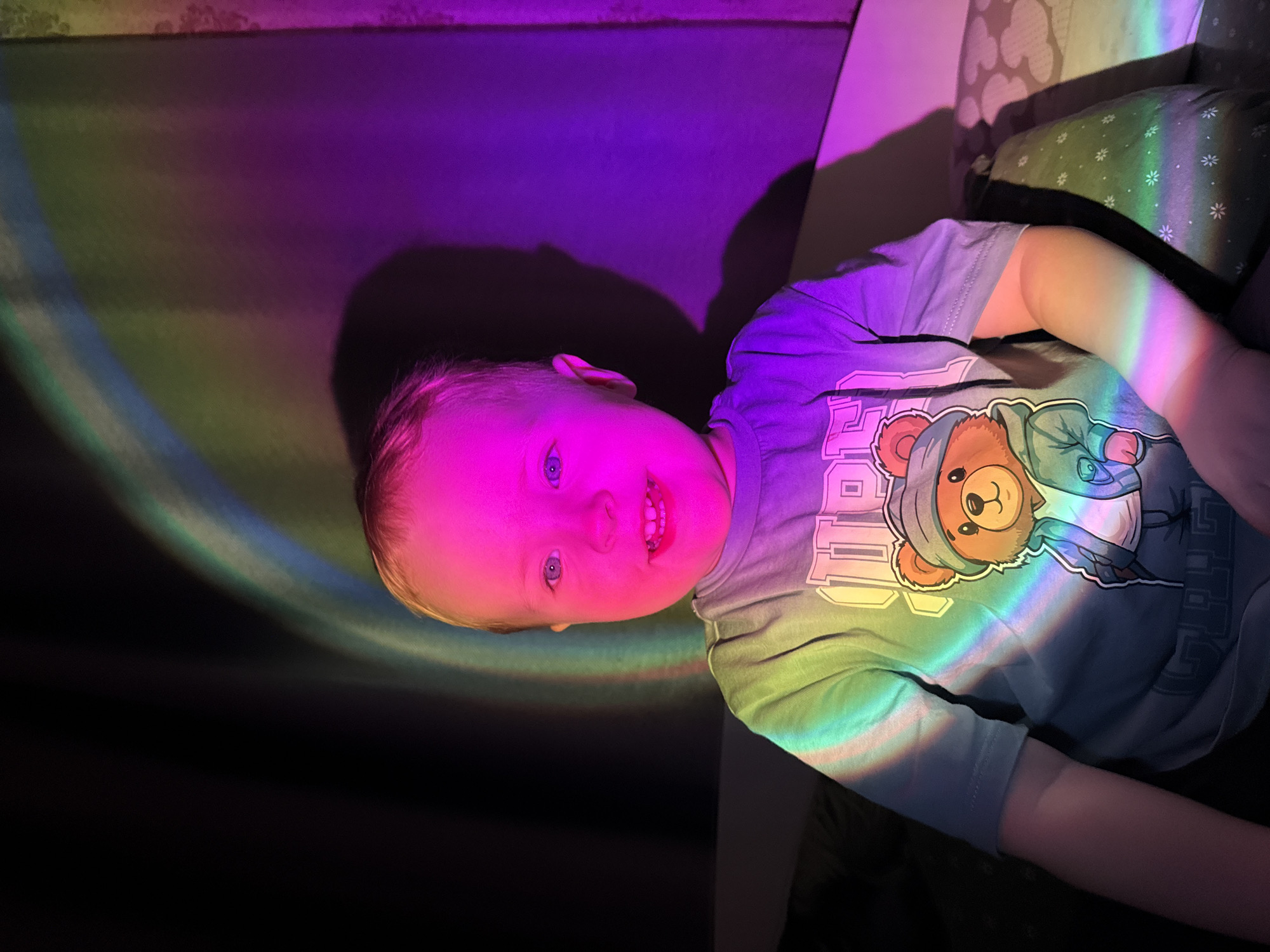An eye-level, close-up shot captures a smiling toddler bathed in a spectrum of colorful light, set indoors in Chaikovsky, Russia. The young child, with light-colored hair and bright blue eyes, is positioned horizontally in the frame, looking directly at the viewer with a joyful, toothy smile. A vibrant rainbow effect, likely from a light projector or prism, illuminates the scene. The child's face is predominantly bathed in bright pink and purple light, while hues of green, yellow, orange, and blue extend across their body and surroundings.
The child is wearing a light blue or gray t-shirt featuring a graphic of a brown bear with a blue hoodie and glasses. Some white text, possibly "P.I.P." or similar, is partially visible on the upper part of the shirt. The colorful light also illuminates the child's arms and a patterned pillow or cushion to their right. A textured wall forms the background, with the child's shadow cast in a deep purple on the left. The overall ambiance is playful and enchanting, emphasizing the child's happy and curious expression.