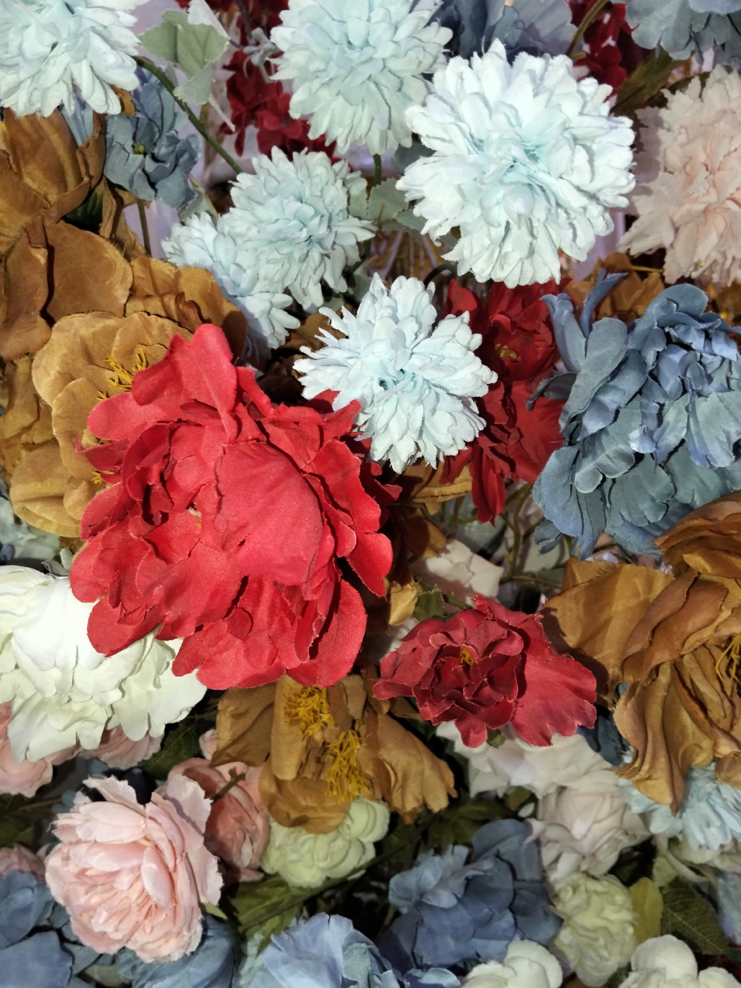 The image is a close-up, high-angle shot of a dense arrangement of artificial flowers. The flowers are varied in color and type, including large red peonies, pale blue chrysanthemums, muted blue dahlias, and pale pink roses. The arrangement also features some dried or faux flowers in shades of brown and cream. The background is a soft, out-of-focus white. The lighting is even and highlights the textures of the fabric flowers. There are no people, actions, or interactions depicted. The scene is likely a floral decoration or display, possibly in an indoor setting. The location of Cottrueng, Indonesia, is not visually evident in the image.