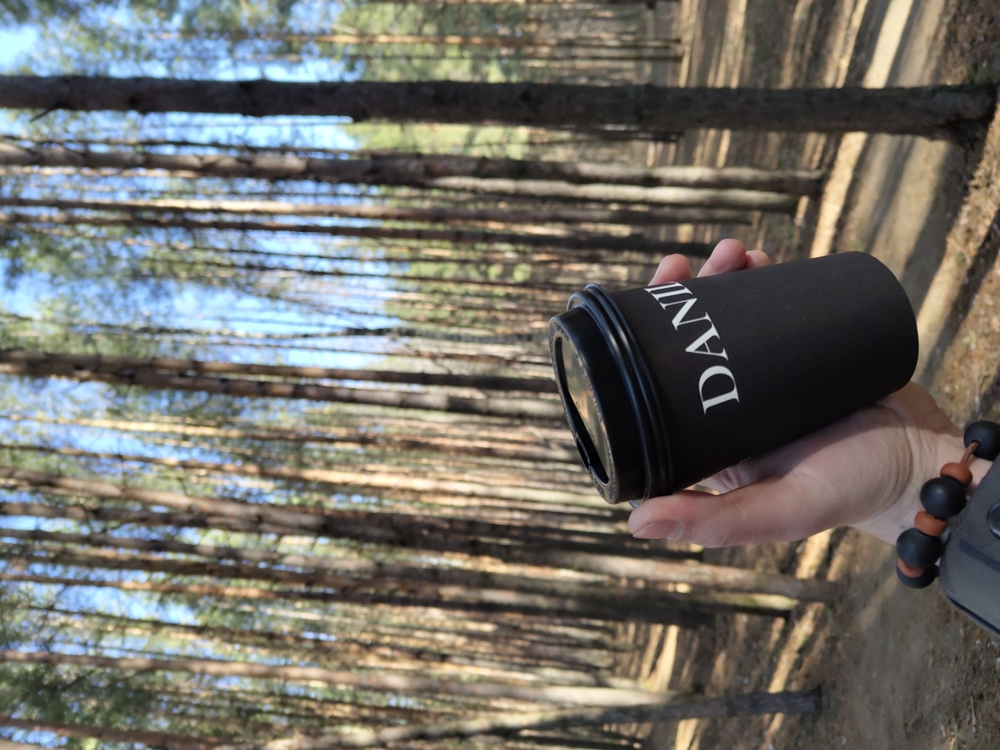 The image shows a hand holding a black cup with the word "DANI" printed on it. The background is a forest with tall trees and a clear blue sky visible through the branches. The location is the city of Chaikovsky, Russia, suggested by the trees and sky. The person is wearing a bracelet made of black and brown beads. The hand is holding the cup at an angle. The trees are casting shadows on the ground.