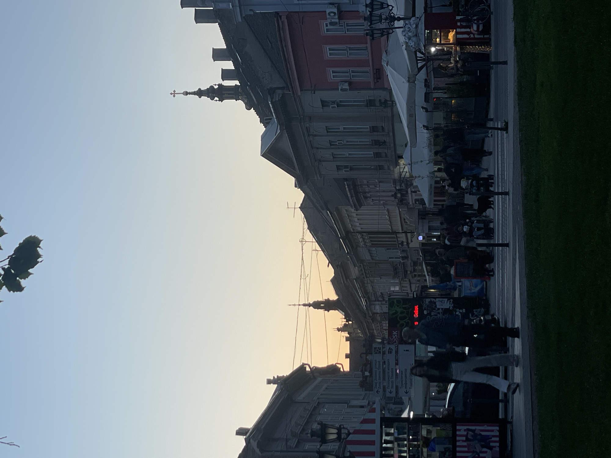 The image captures a street scene in Novi Sad, Serbia, oriented vertically. Buildings line either side of the street, diminishing into the distance towards the horizon. The architecture varies, featuring multiple stories and distinct rooflines, some topped with steeple-like structures. Pedestrians populate the street, with figures walking and gathered near outdoor cafe seating. The sky is light, with a gradient suggesting either sunrise or sunset. Street furniture, including umbrellas and street lights, lines the pedestrian area. A sign displaying street names and tourist information is visible on the right.