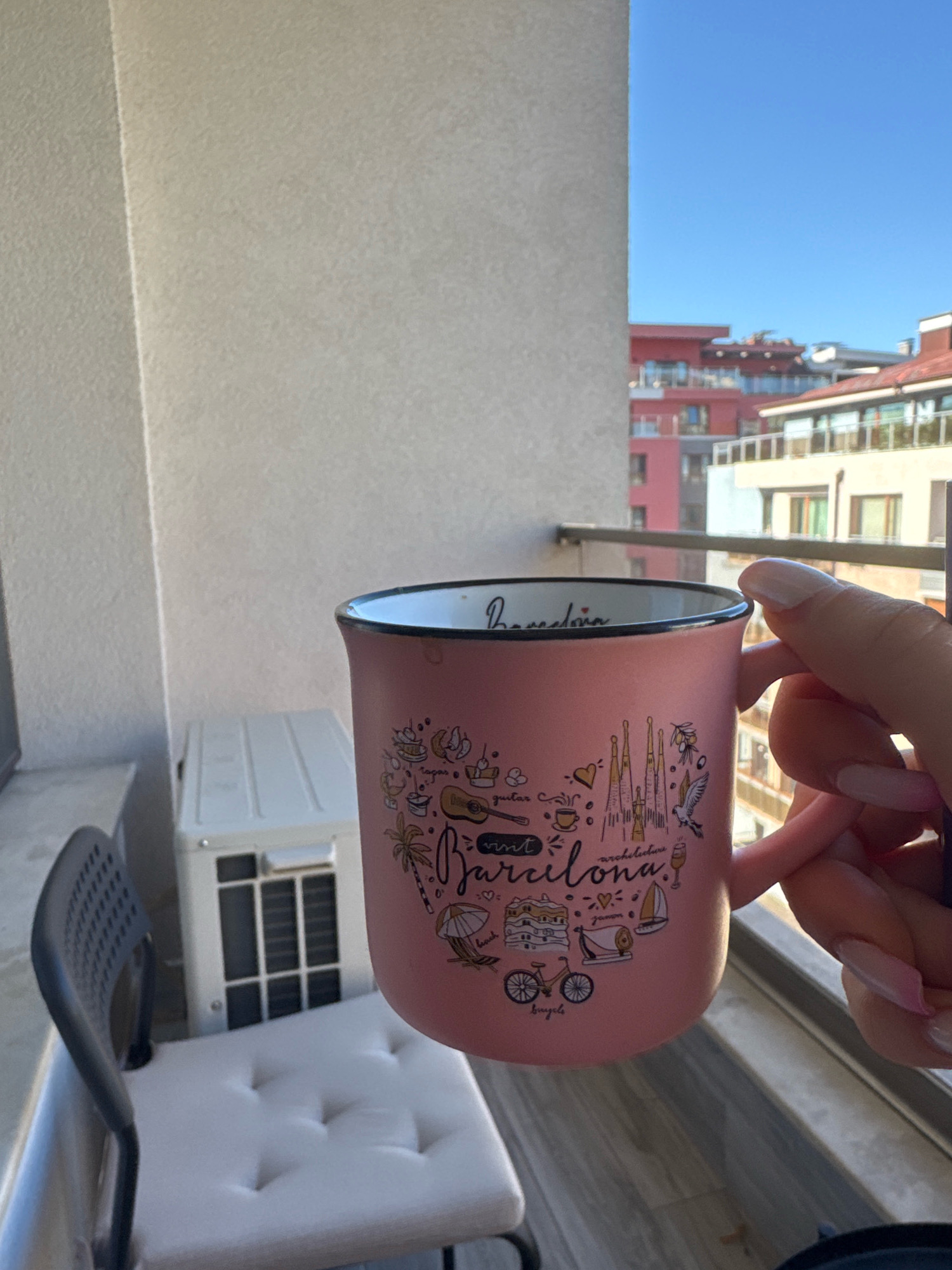A person's hand with manicured nails holds a pink mug with "Barcelona" written on it, surrounded by various icons representing the city, including Sagrada Familia, a guitar, a bike, and a sailboat. The mug rests on a balcony railing, with a view of apartment buildings and a clear blue sky in the background. An air conditioning unit and a chair with a tufted cushion are also visible on the balcony. The scene is brightly lit by sunlight.