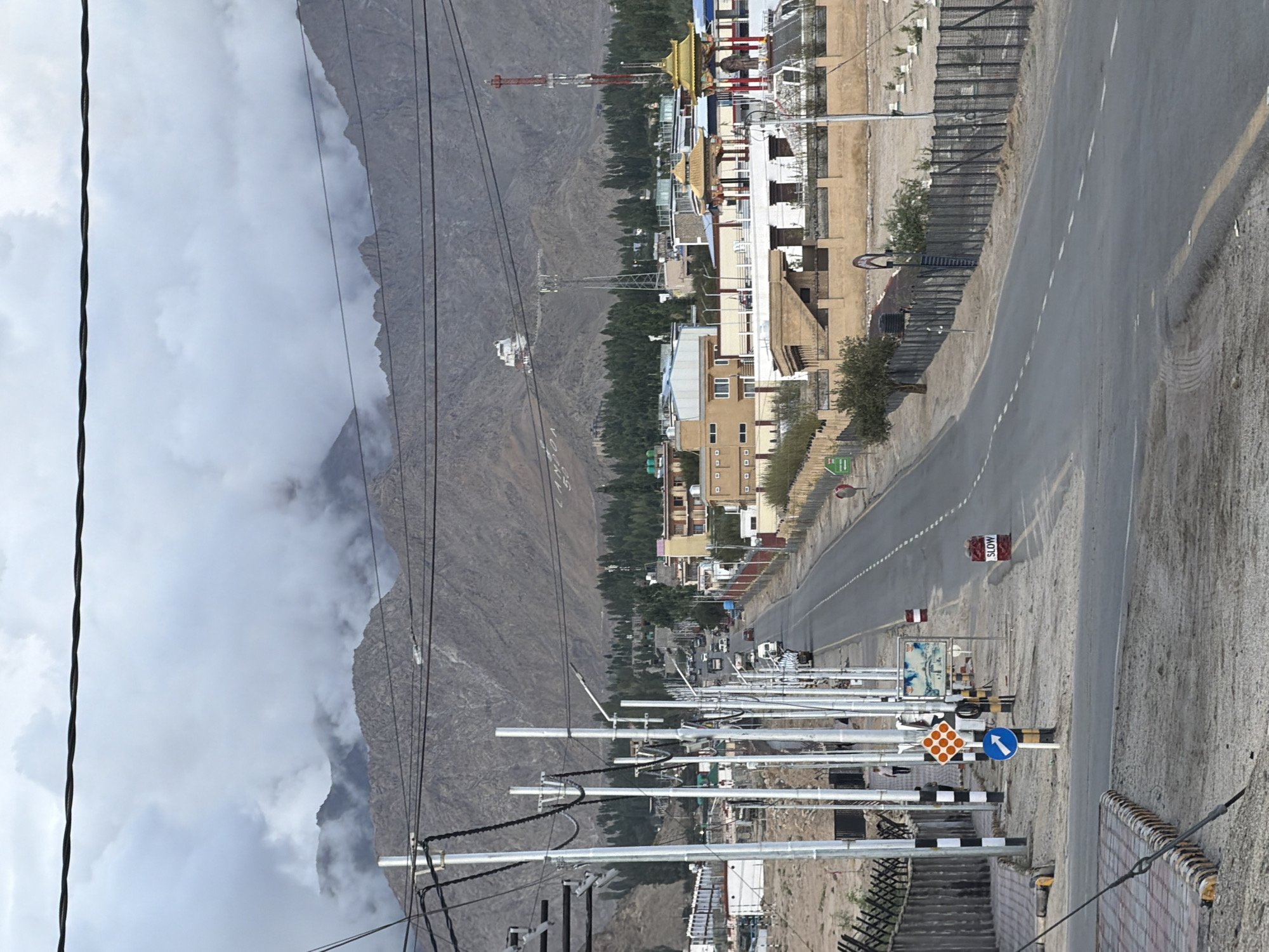 The image shows a wide, paved road leading into a town nestled at the base of barren, mountainous terrain. The sky is overcast with heavy clouds, obscuring the peaks of the mountains in the distance. Along the road, there are numerous utility poles with electrical wires crisscrossing overhead. The town exhibits a mix of modern and traditional architecture, with multi-story buildings and some structures featuring golden-roofed pavilions. A tall red and white telecommunications tower is visible in the middle ground. Several vehicles, including cars and a truck, are driving on the road, indicating ongoing activity. Traffic signs, including a "SLOW" sign and directional arrows, are present. The roadside features a fence, some sparse vegetation, and pedestrian pathways. The overall impression is of a developed area situated in a rugged, high-altitude environment. The context of Spituk, India suggests this is likely a view of the road approaching or within the town of Spituk, a significant Buddhist center in Ladakh.