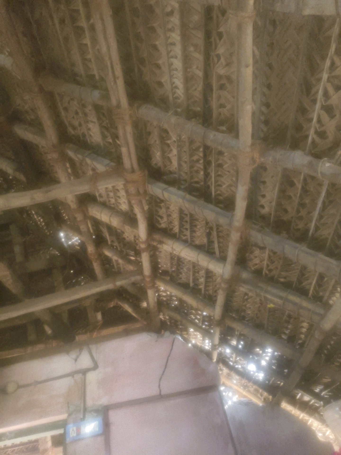 This image shows the underside of a thatched roof, likely in Therkkusalli kulam, India. The roof is constructed from palm fronds or similar vegetation, supported by a framework of wooden beams and poles. The beams are held together with twine or rope. Sunlight filters through the gaps in the thatch, creating bright spots and shadows. In the lower part of the image, a section of a wall is visible, with electrical conduit and a junction box or light switch. The overall scene suggests a rustic or traditional building, possibly a dwelling or a storage area, in a rural or semi-rural setting.