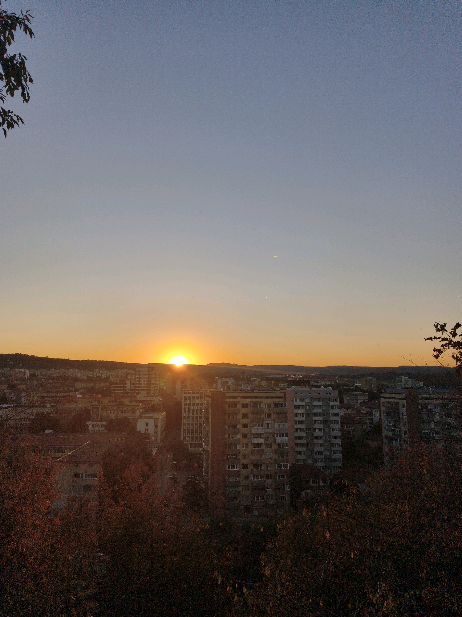 The image captures a sunset over the city of Lovech, Bulgaria. The sun is a bright orb, just above the horizon, casting a warm golden glow across the sky and the city below. The city appears to be comprised of apartment buildings of varying heights, with some residential structures and what may be commercial buildings visible. Trees with autumnal foliage are in the foreground, partially obscuring the view of the city, suggesting the season is autumn. The sky is a gradient of soft blue and orange hues, with the setting sun creating a vibrant transition. The overall scene is one of peaceful urban twilight.