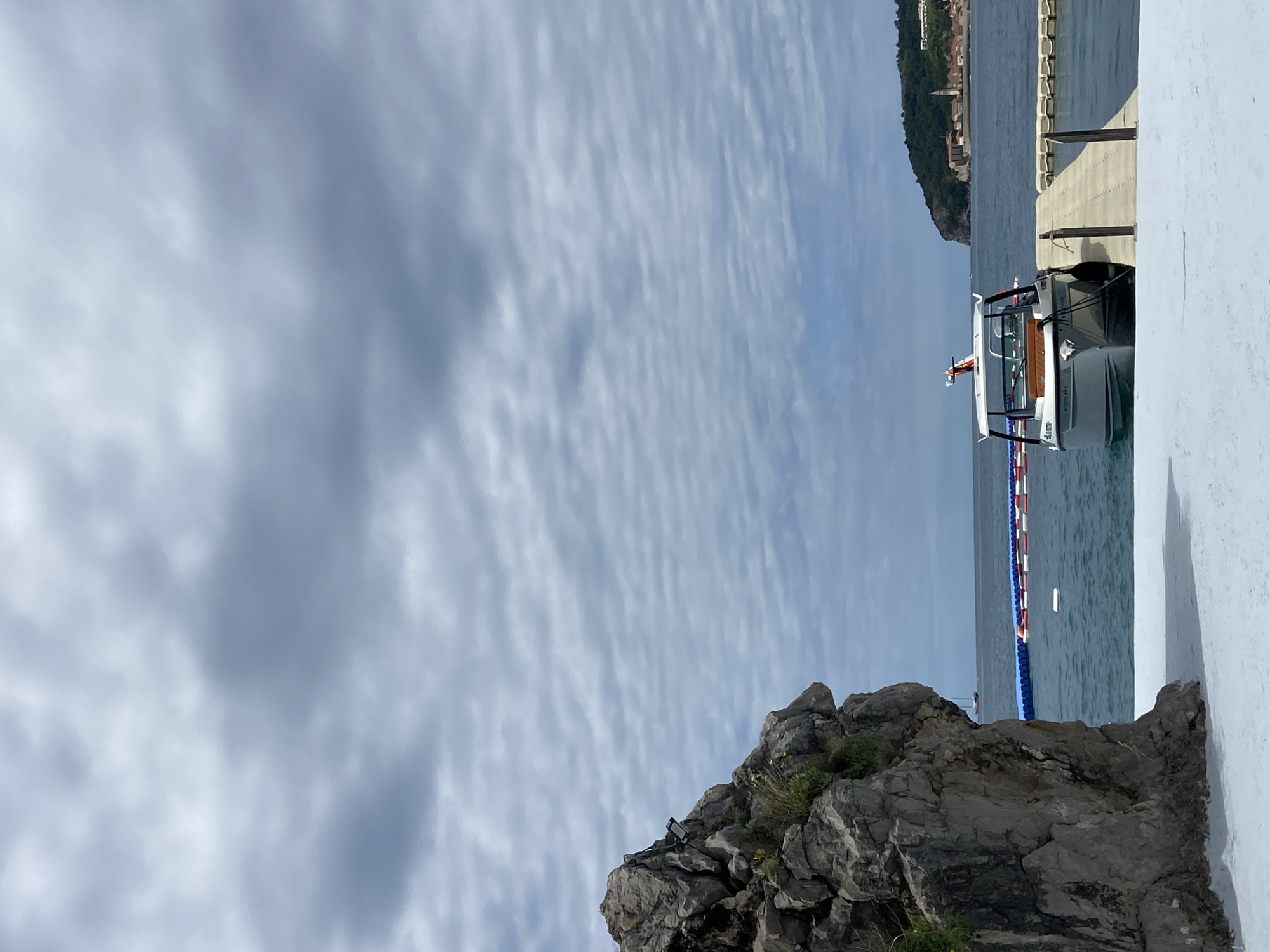 The image shows a scene at the harbor of Budva, Montenegro. The sky is overcast with stratocumulus clouds. A rocky outcrop with sparse vegetation is located in the foreground. To the right of the rocks, a white building structure is visible. The harbor features a pier with a boat docked alongside. A string of red, white, and blue buoys is present in the water. In the distance, a hill with buildings on top is discernible.