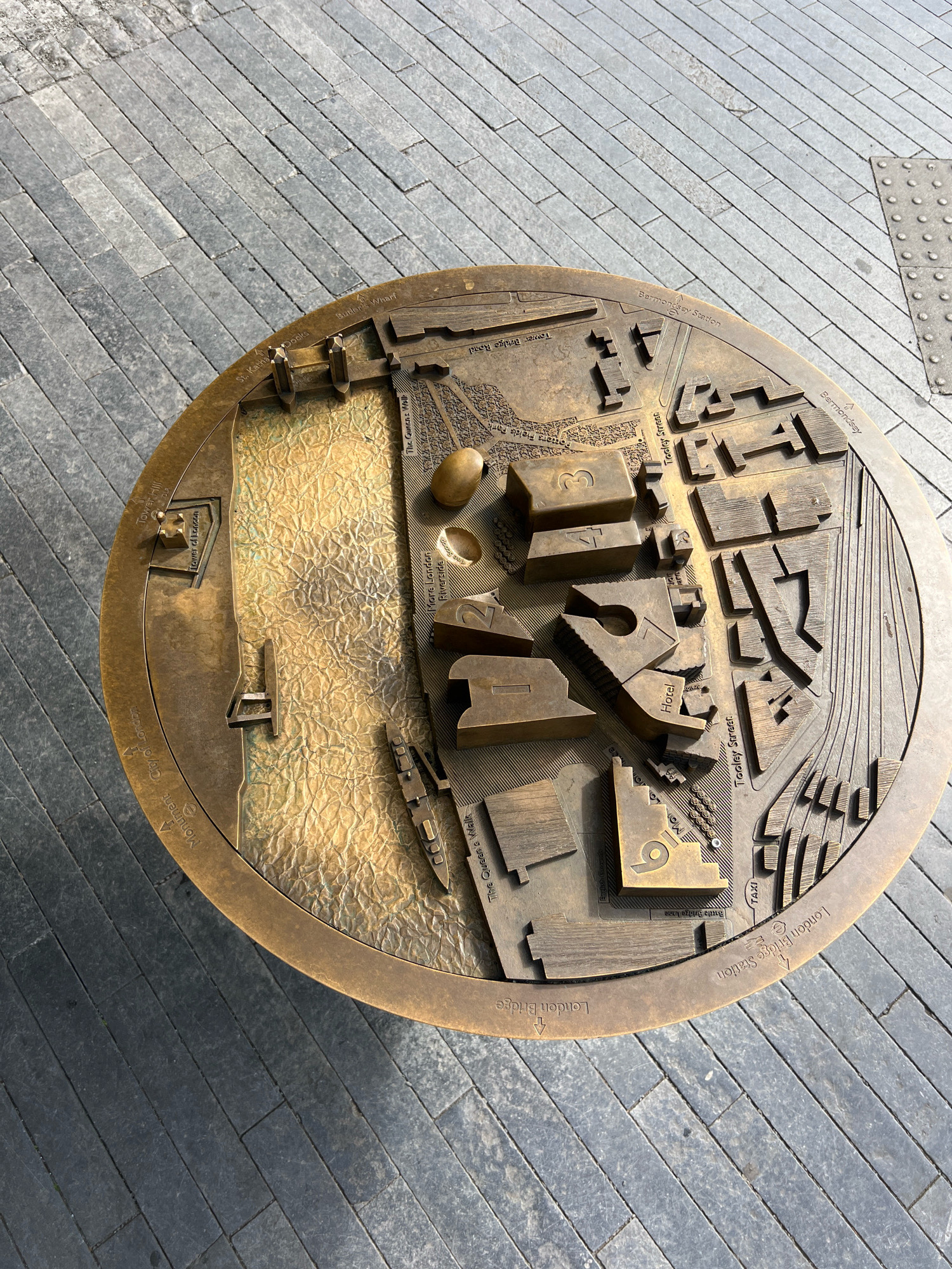 This is a top-down, slightly angled, medium shot of a bronze relief map situated on a grey, paved sidewalk. The map depicts an area of London, United Kingdom, with raised structures representing buildings and textured areas indicating water and terrain. Key landmarks and street names are engraved on the map's surface, including "Tower of London," "London Bridge Station," "Tower Bridge Road," "Bermondsey," and "St. Katharine Docks." A portion of the River Thames is also represented with a textured, crinkled golden surface. The map is circular in shape and is embedded in a sidewalk paved with dark grey rectangular stones laid in a herringbone pattern. The lighting suggests a sunny day, casting shadows from the raised elements of the map. There are no people or animals visible in the image. The environment is an outdoor urban setting.