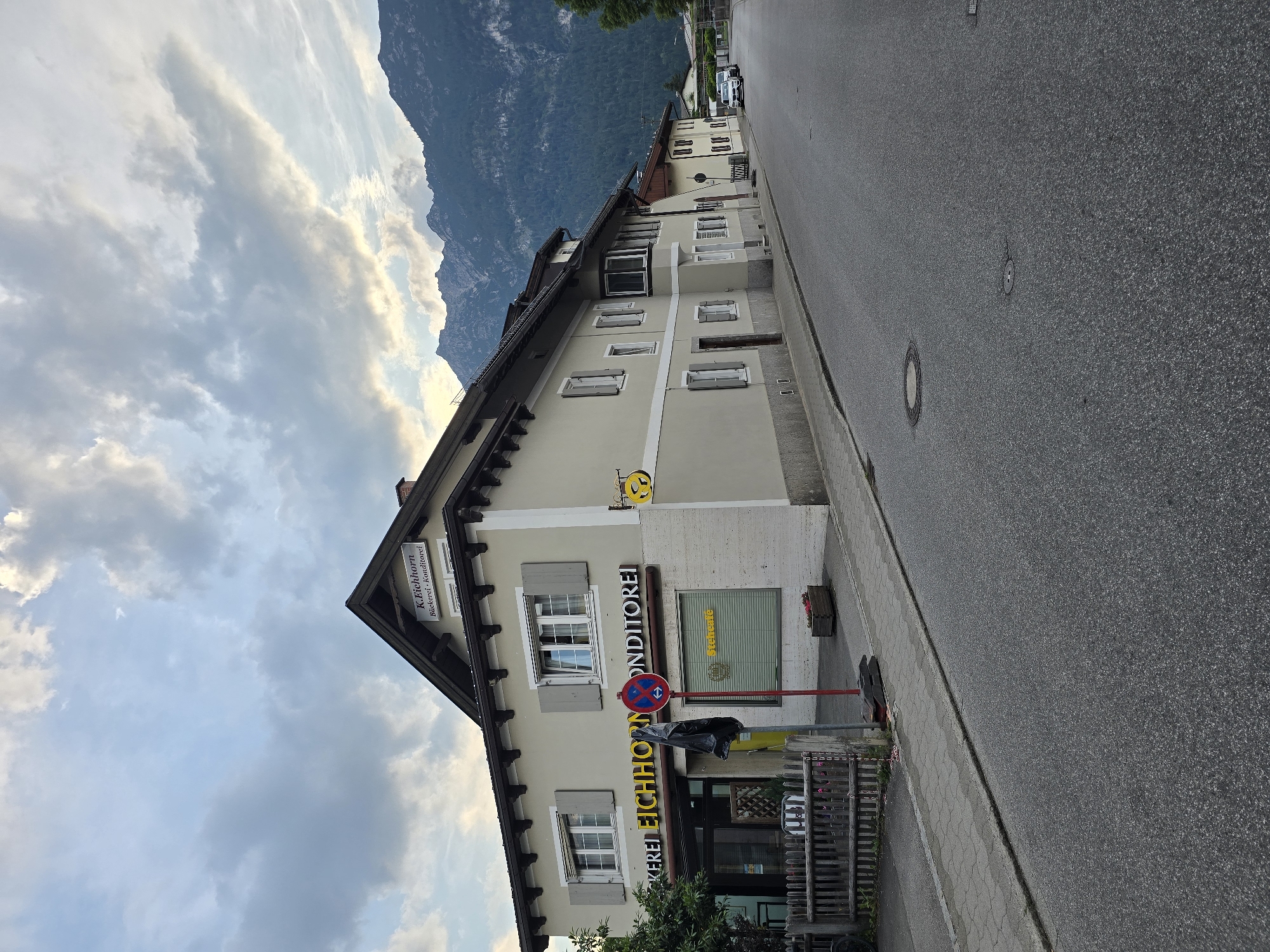 The image shows a street in Garmisch-Partenkirchen, Germany, with a building on the left side of the road. The building appears to be a bakery and confectionery shop named "Bäckerei Eichhorn Konditorei". The shop has large windows and signs indicating its business. In front of the shop, there is a wooden fence and a red and white sign. The street is paved and has a sidewalk. In the background, there are mountains covered in trees, and the sky is partly cloudy. There are cars parked on the right side of the street further down. The overall scene depicts a quiet street in a town with a mountainous backdrop.