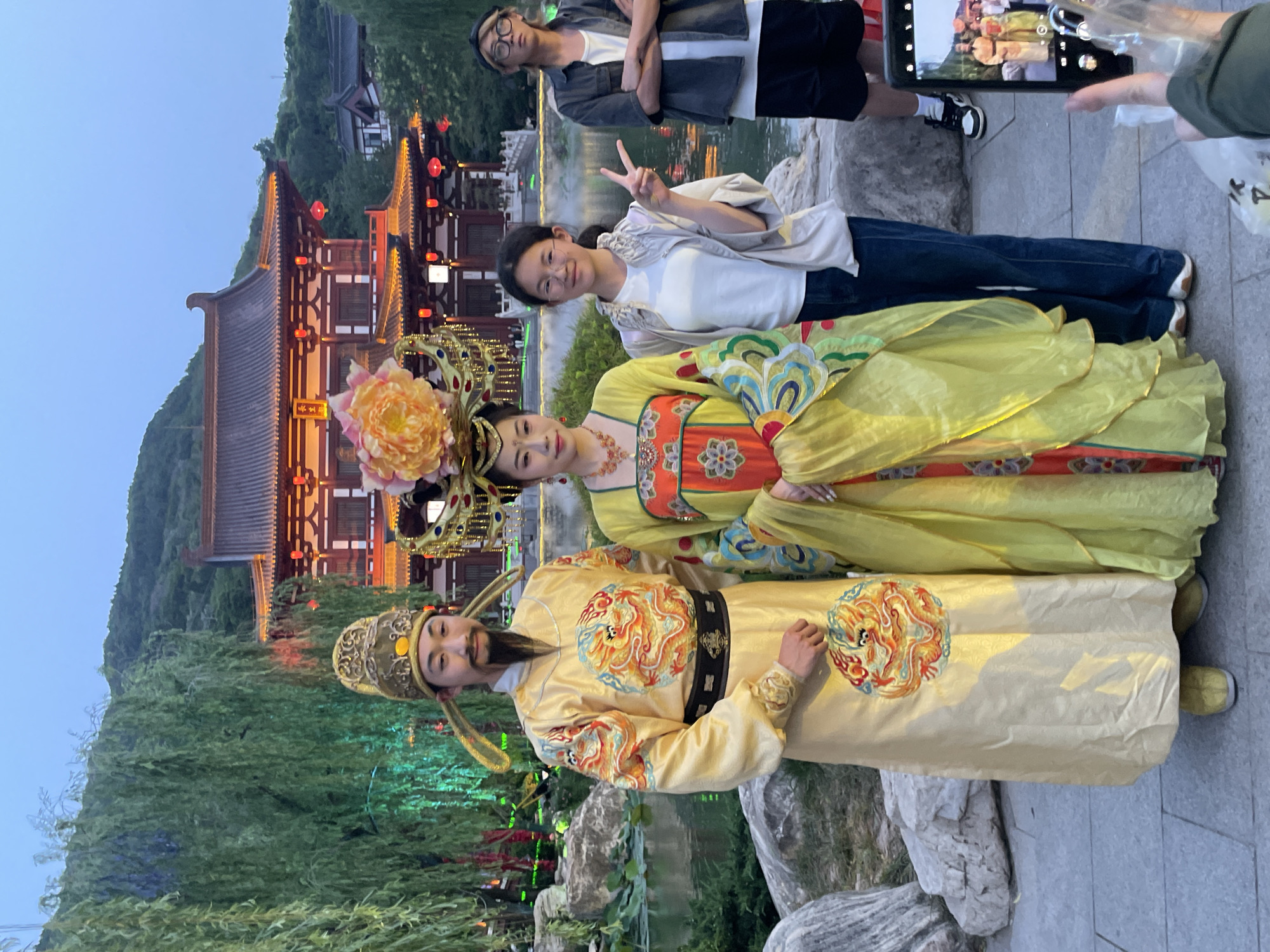This nighttime image depicts a group of people posing for a photo in Lintong District, China. In the foreground are two individuals in traditional Chinese attire, one dressed as a man with a beard and ornate headdress, and the other as a woman with elaborate floral headwear. Behind them, two younger individuals are visible, one holding up a peace sign and the other presumably taking the photo with a mobile phone. The backdrop features traditional Chinese architecture with illuminated red lanterns, nestled against a hillside with greenery. The setting appears to be a garden or park-like area with decorative rocks and water features.
