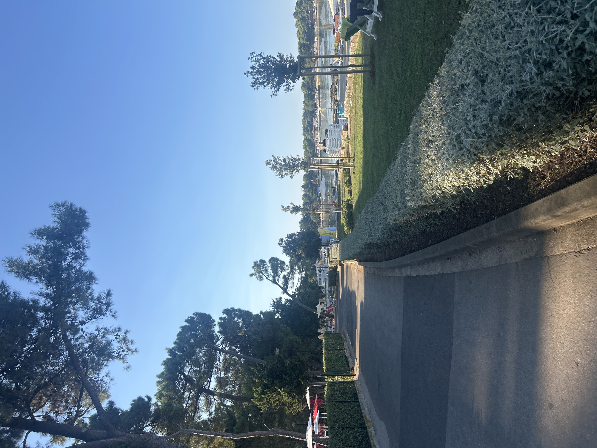 The image depicts a paved walkway running alongside a manicured hedge, with a grassy area beyond. In the background, a beach scene unfolds with numerous lounge chairs, umbrellas, and a body of water dotted with boats. Trees, primarily pines, line the left side of the walkway, casting shadows on the path. The sky is clear and blue, indicating fair weather. The scene is located in Monterol, Croatia, a coastal town. The time appears to be late morning or early afternoon due to the angle of the sun. No specific individuals or actions are discernible in detail, but the presence of beach amenities suggests a leisure or resort area.