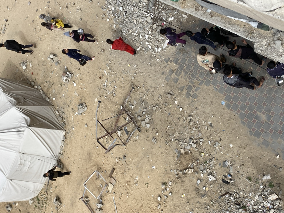 An overhead view of an outdoor area in Khan Yunis, Palestinian Territory, depicts a scene characterized by sandy ground, scattered rubble, and multiple individuals. In the upper left, a large, light-colored hexagonal tent structure stands on sandy terrain. To its right, an adult male in dark clothing walks away from the tent. Below him, four children, two of whom carry silver metal bowls, traverse the sandy ground from left to right. Further down, an individual in a red tracksuit walks rightward along the edge of a rubble-strewn patch. Prominently in the central left, two twisted, open-frame metal structures, possibly from damaged furniture, lie on the sand amidst debris. In the lower right, a group of six individuals, including adults and older children, walk along a patterned paved walkway bordered by a concrete barrier on the far right. At least three individuals in this group are also carrying silver metal bowls. The ground throughout the unpaved areas is a mix of light-brown sand and numerous pieces of concrete, stones, and general debris.