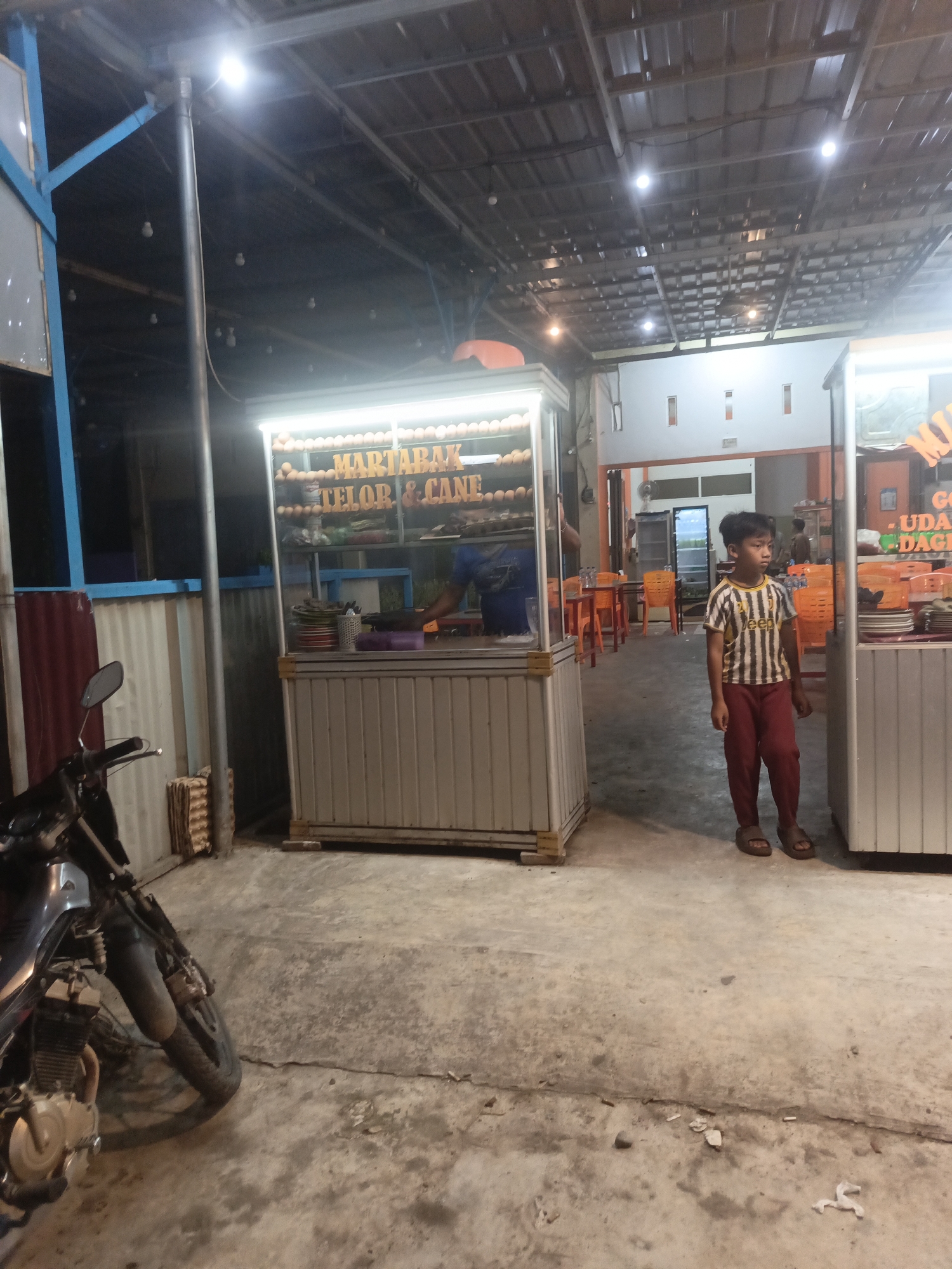 This image depicts an outdoor food stall, likely in Lhokseumawe, Indonesia, at night. A young boy wearing a striped t-shirt and red pants stands in front of a food cart. The cart has a display of eggs at the top and appears to be selling "MARTABAK TELOR & CANE." Behind the boy, there are orange plastic chairs and tables, suggesting a dining area. To the left of the frame, a motorcycle is parked. The environment appears to be a covered outdoor space with metal roofing and lighting.
