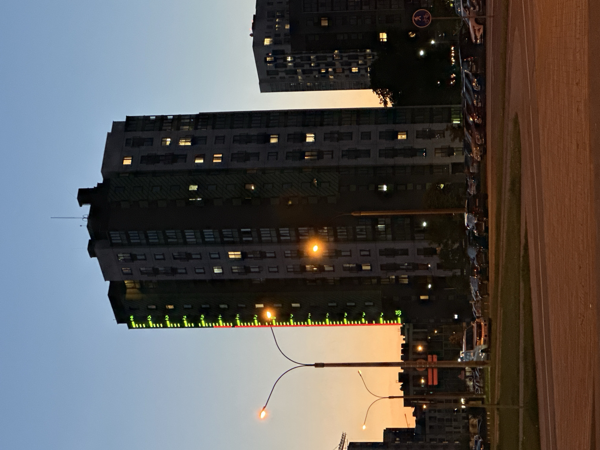 The image captures an urban or suburban street scene at dusk in Бараўлянскі сельскі Савет, Belarus. Multiple tall, dark buildings with numerous illuminated windows dominate the frame, suggesting residential or office use. The sky transitions from a soft blue at the top to warm orange and yellow hues near the horizon, characteristic of twilight.
A prominent multi-story building stands in the center, featuring a distinctive long, horizontal digital display along one of its lower levels. This display shows a series of green and red vertical bars, resembling a large outdoor thermometer or a dynamic information board. Visible numbers on this display include "+3", "+5", "-3", and "35", separated by small dots and tick marks, with a continuous red line running horizontally through its center.
Several streetlights with bright orange-yellow glows are scattered throughout the scene, illuminating the surroundings. A prominent 'Y'-shaped streetlight fixture is visible in the mid-ground. To the right, a multi-lane road or highway is partially visible, featuring a grassy median. A line of parked cars can be seen along the road or an adjacent parking area. A blue circular sign with a white bicycle symbol, indicating a bicycle path or lane, is visible near the top-right of the main building. Dark foliage from trees and bushes is silhouetted against the sky and building lights, adding to the evening ambiance. The scene is static, depicting the calm transition from day to night in a developed area.