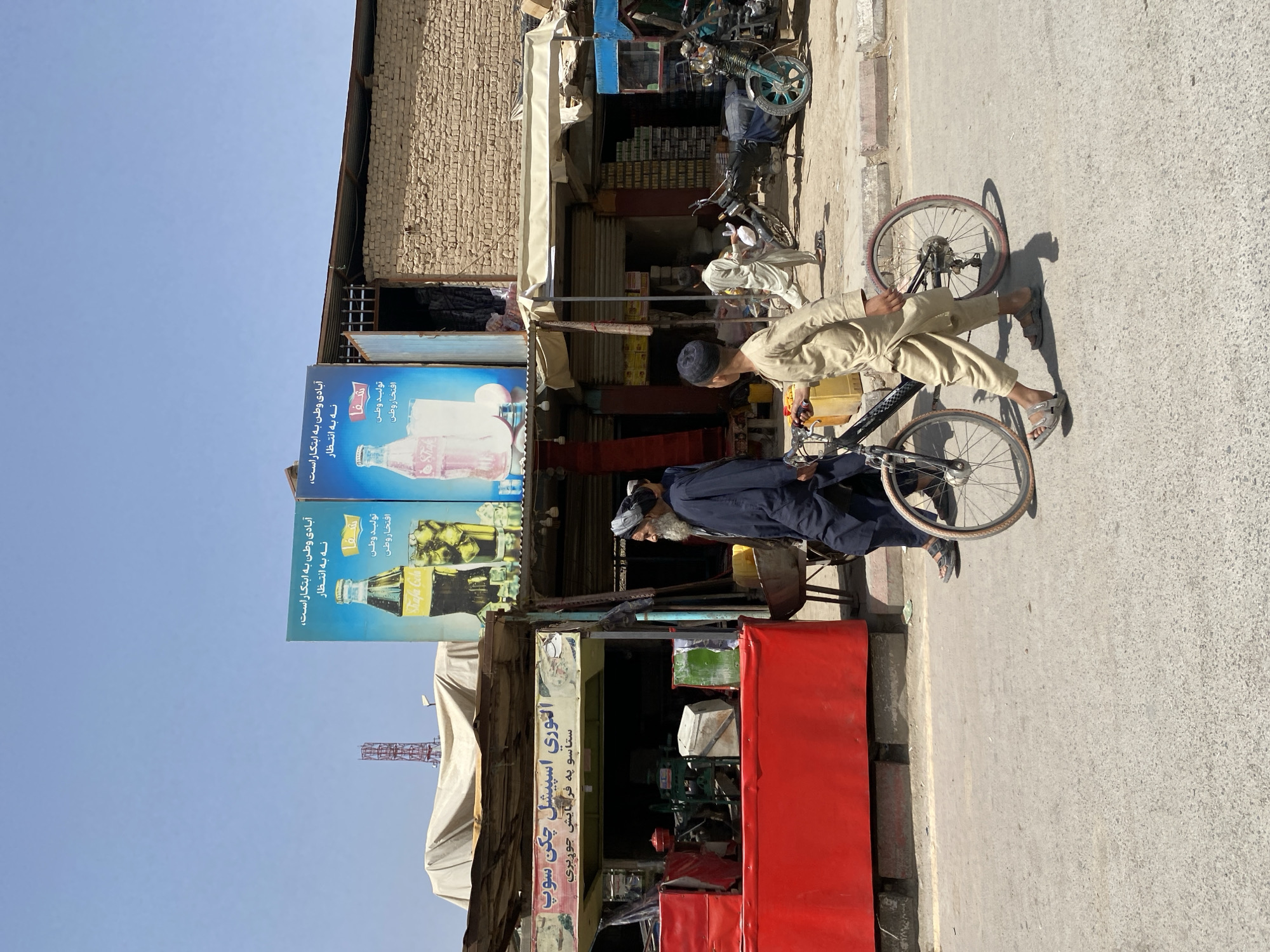 The image depicts a street scene in Kandahar, Afghanistan. The foreground shows a paved road with a boy walking his bicycle. The boy is wearing a tan outfit and sandals. Behind him, an older man in a dark blue robe and turban walks in the same direction. To the left of the road is a market stall with a bright red counter, displaying various items and a red generator. Above the market stall, an advertisement for "Shifa Cola" is visible. Further down the street, a brick building with an open doorway is seen. In front of the building, a man in a light-colored robe is walking. Various motorcycles and a small cart are parked along the street. The sky is clear blue.