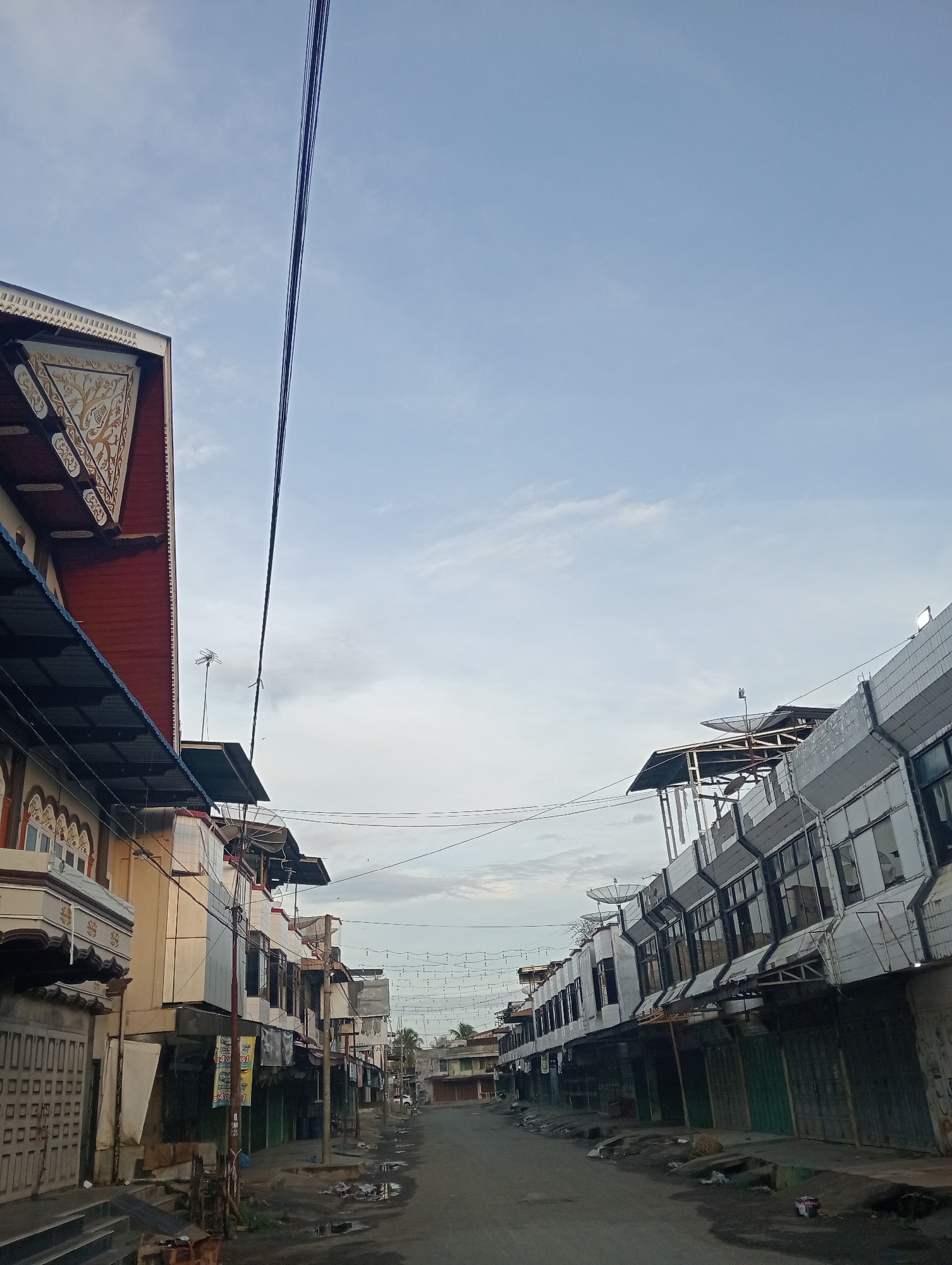The image shows a street scene in Keudegeudong, Indonesia, with buildings on both sides of the road. The sky is partly cloudy and a light blue. Overhead, power lines run across the scene, with one thick black cable dominating the left side of the frame.
On the left side of the street, there is a building with a red roof and ornate white trim. The building has several windows and a covered walkway. Further down the street on the left, there are more buildings with similar architectural styles.
On the right side of the street, there is a row of commercial buildings with modern facades. These buildings appear to be shops or offices, with shutters or closed doors visible. Satellite dishes and antennas are mounted on the roofs of some of these buildings.
The road itself appears to be paved and wet, with puddles of water reflecting the sky and surrounding buildings. There are some discarded items and debris on the side of the road. The scene suggests a quiet, possibly early morning or late afternoon, with minimal human activity visible.