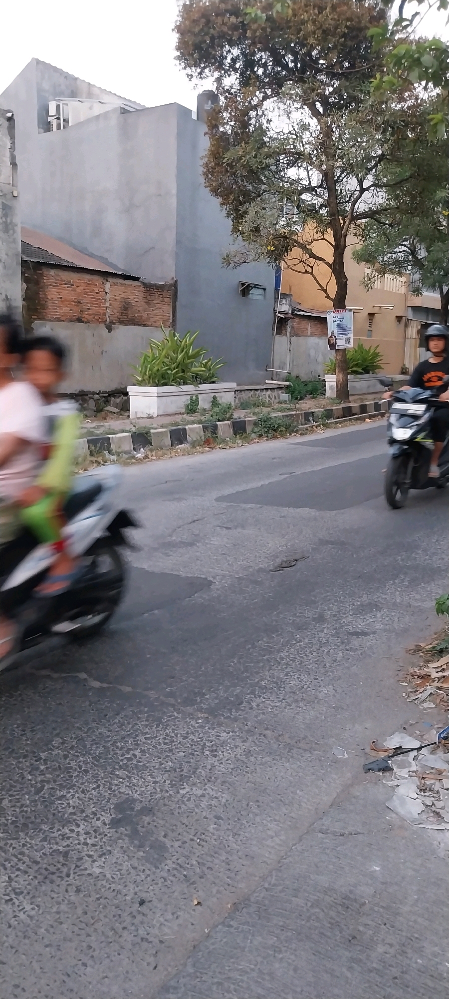 The image shows a street scene in Bojong Nangka, Indonesia. A motorcycle carrying two people is in motion on the left side of the frame, with the passengers blurred due to the movement. On the right side of the road, another motorcycle with a single rider wearing a helmet is also moving. Buildings with gray and brick exteriors are visible in the background, along with trees and vegetation. A planter with green plants is situated by the roadside. The road surface appears to be asphalt with some wear and tear. The overall lighting suggests it is daytime, possibly late afternoon.