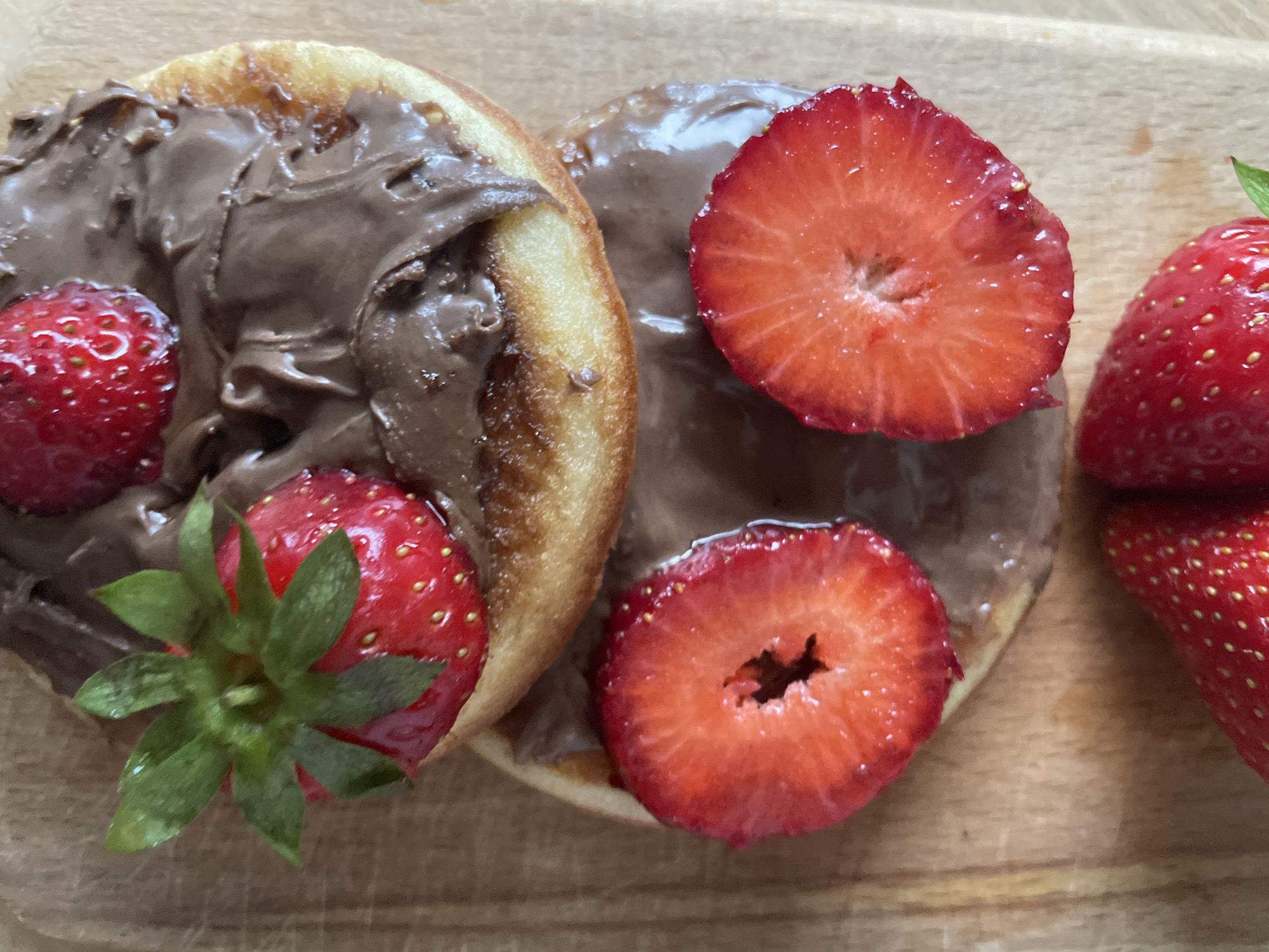 The image displays a close-up of two round pastries, possibly pancakes or small donuts, generously topped with a thick, dark brown chocolate or hazelnut spread. On the left pastry, a vibrant red whole strawberry with its green calyx (stem and leaves) is placed, along with another whole strawberry partially visible near the top left. A cut half-strawberry, revealing its bright red pulp and internal structure, also rests on this pastry. The right pastry features two more halves of strawberries, similarly showcasing their juicy interior and tiny seeds. Additional whole strawberries are visible on the far right of the frame, partially cropped. All the food items are arranged on a light brown wooden surface, likely a table or cutting board, suggesting a prepared breakfast or dessert. The natural, soft lighting indicates a daytime setting. No text is visible in the image. The context for this scene is Plovdiv, Bulgaria.