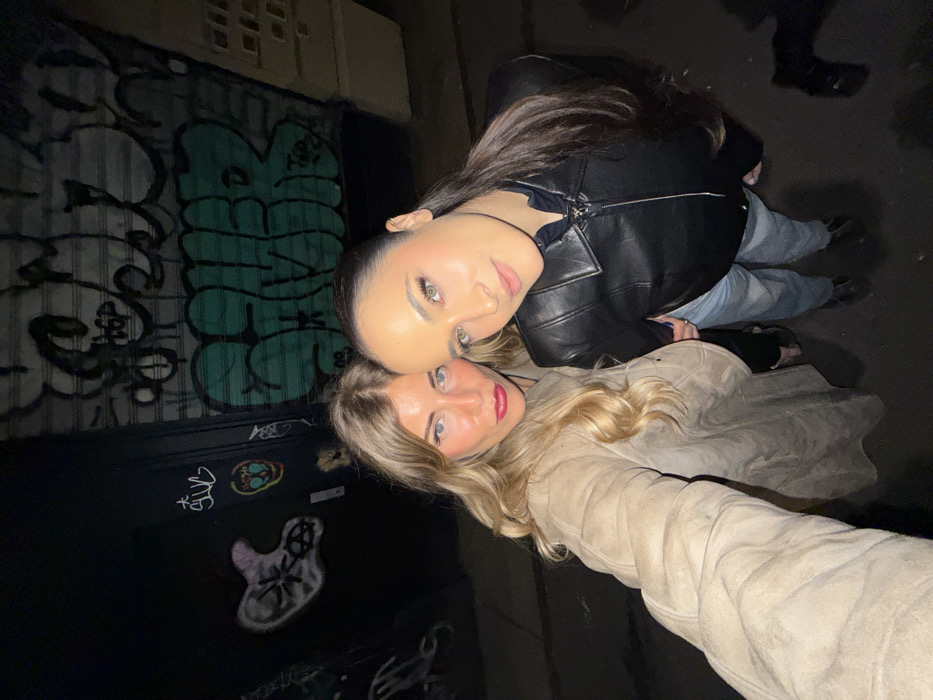 A street-level, low-light photograph taken in the city of Paris, France, depicts two adult females posing closely for a selfie. The woman on the left, with long blonde hair and bright red lipstick, wears a light beige or tan long coat. She extends her right arm, holding the camera, and looks directly forward. Adjacent to her, the woman on the right has long, dark brown hair pulled into a ponytail. She wears a black leather jacket over a dark top, light blue jeans, and dark shoes, with her head slightly tilted and a subtle smile directed towards the camera. The background on the left features a dark wall marked with various white spray paint graffiti tags and an abstract skull-like image. On the right, a metal rolling security shutter, characteristic of a closed storefront, is extensively covered in green and black spray paint graffiti, including large block letters. The ground beneath the subjects is dark asphalt. The scene is illuminated by an artificial light source, highlighting the subjects' faces against the dim urban environment.