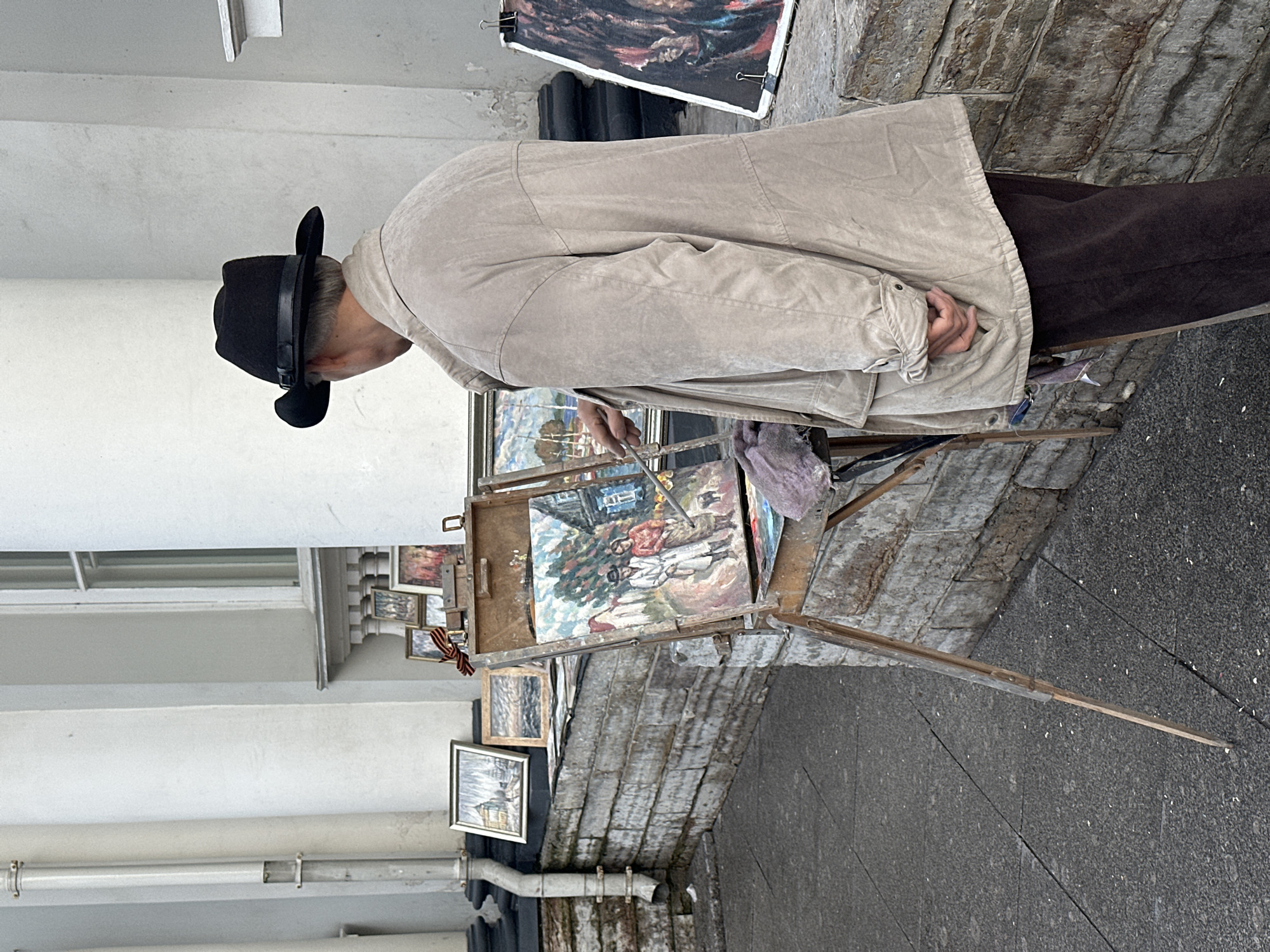 An artist in a black fedora and a beige jacket is painting an outdoor scene in Saint Petersburg, Russia. He stands behind an easel displaying a finished painting of two people and a building. Several other paintings are displayed on a stone ledge behind him, and a red and black ribbon is attached to a wooden box. The scene is set against a white building with classical architectural elements, including columns and window frames. The ground is paved with dark grey cobblestones.