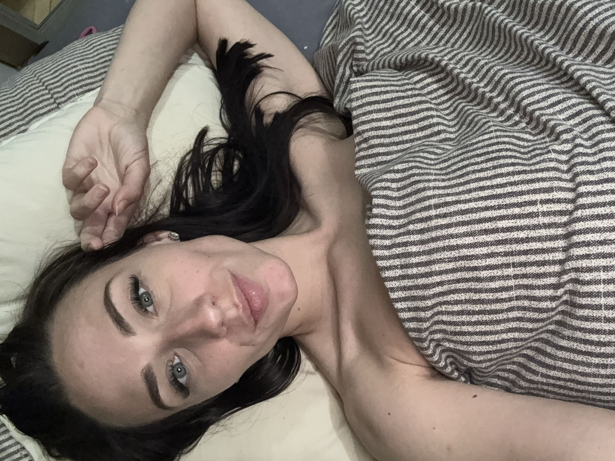 A person with long, dark hair lies on a striped bed under a striped blanket. The person is fair-skinned and has visible eyelashes and eyebrows. Their left arm is raised above their head, with their hand resting on a pillow. A small earring is visible in their ear. The background includes a gray headboard. There is no indication this image was taken in Saint Petersburg, Russia.