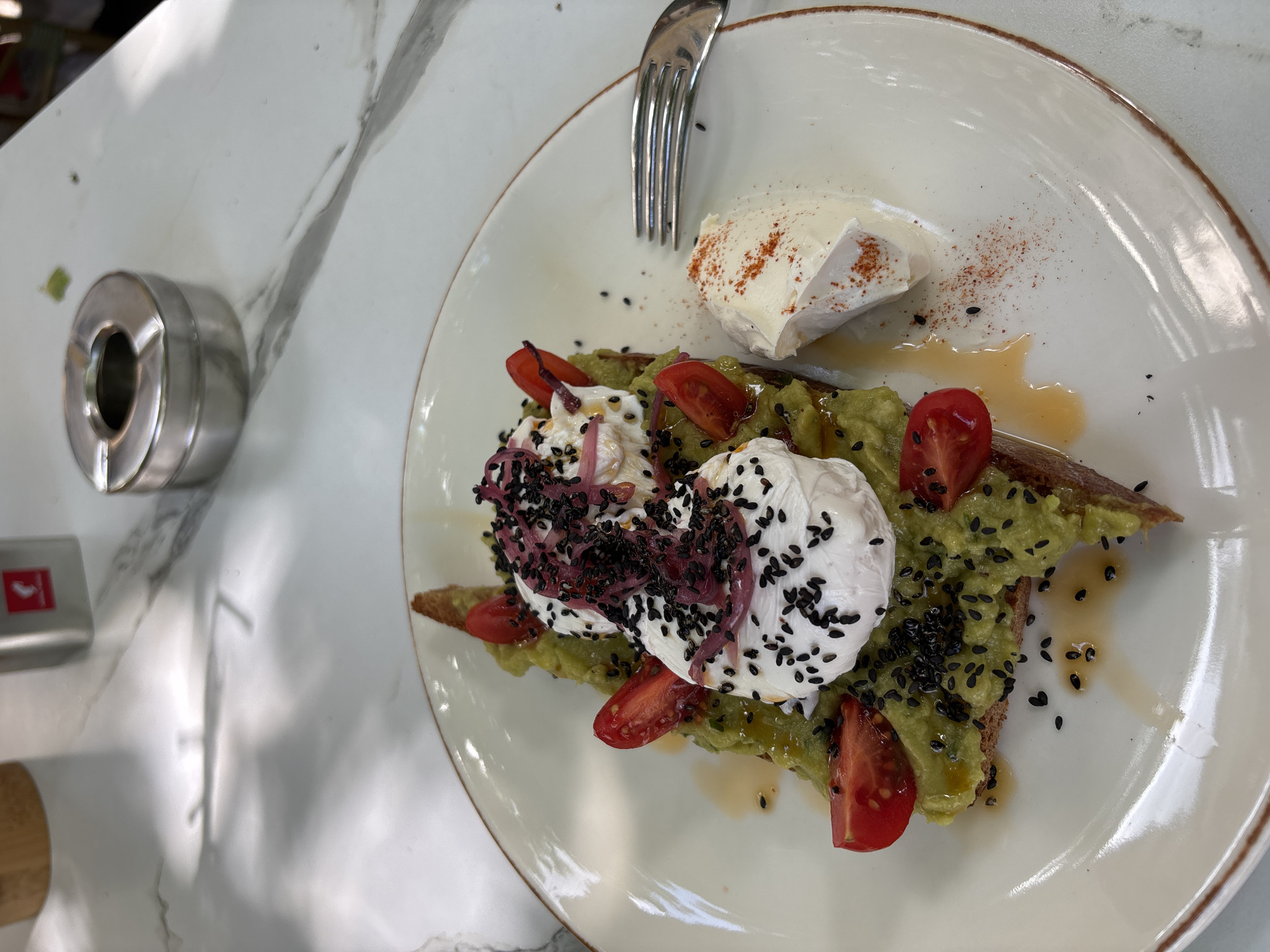 A plate of avocado toast with two poached eggs, halved cherry tomatoes, sliced red onion, and black sesame seeds is presented on a marble-patterned tabletop, possibly in Varna, Bulgaria. A fork rests to the right of the plate, alongside a dollop of cream cheese sprinkled with paprika. In the background, a metallic ashtray and a cylindrical dispenser are visible. The lighting suggests an outdoor or well-lit indoor setting.
