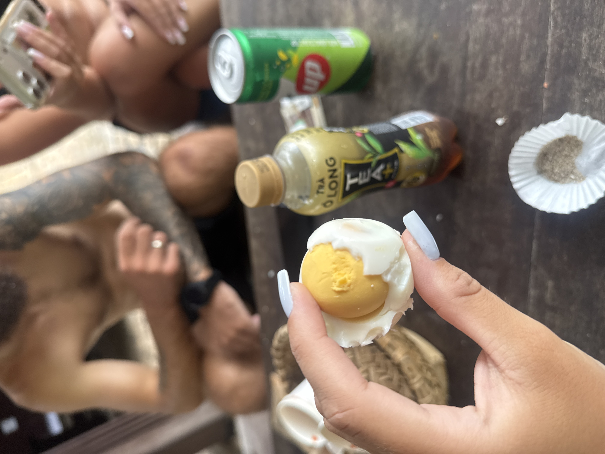 In Nha Trang, Vietnam, on a wooden table, a person's hand with light blue nails holds a half-peeled hard-boiled egg. The egg has a bright yellow yolk and a white albumen. Beside the egg, a bottle of "Tra O Long Tea+" is visible. In the background, two people are seated. One person, a male, is shirtless and appears to be wearing a ring. The other person, whose body is partially visible, is holding a smartphone. A green can of "Up" soda is also on the table. The setting appears to be an outdoor or casual environment, possibly a beachside cafe or restaurant, given the wicker object behind the egg, which resembles a basket or placemat.