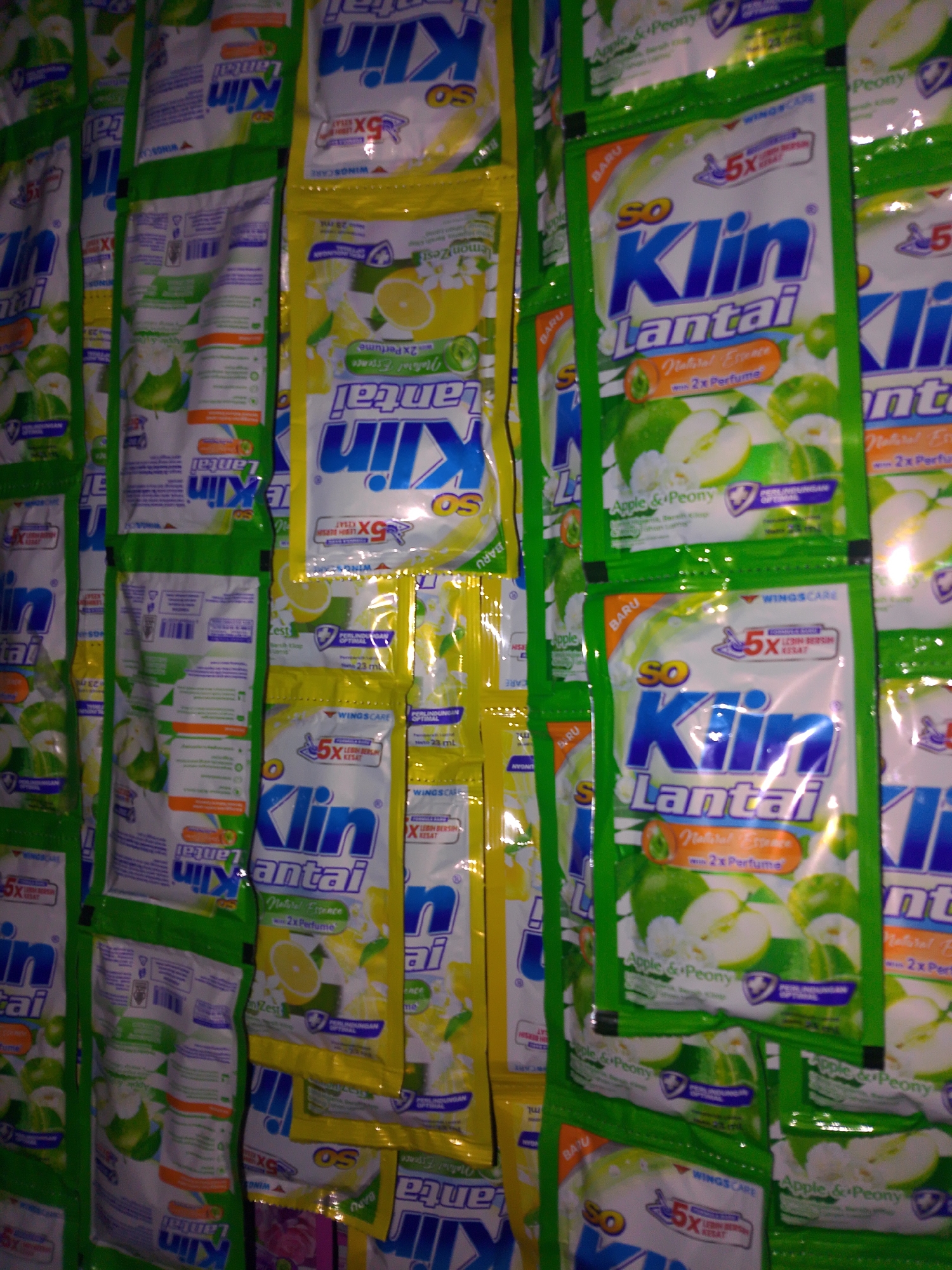 The image displays a dense arrangement of small, individually packaged sachets of floor cleaner, predominantly featuring the "So Klin Lantai" brand. The sachets are predominantly green and yellow, with some white and blue accents, and depict images of apples, lemons, and flowers, suggesting different scents. The arrangement suggests a retail display, likely in a market or small store in Palembang, Indonesia. There are no people or discernible actions visible in the image. The background is completely filled with these products, creating a wall of packaging.