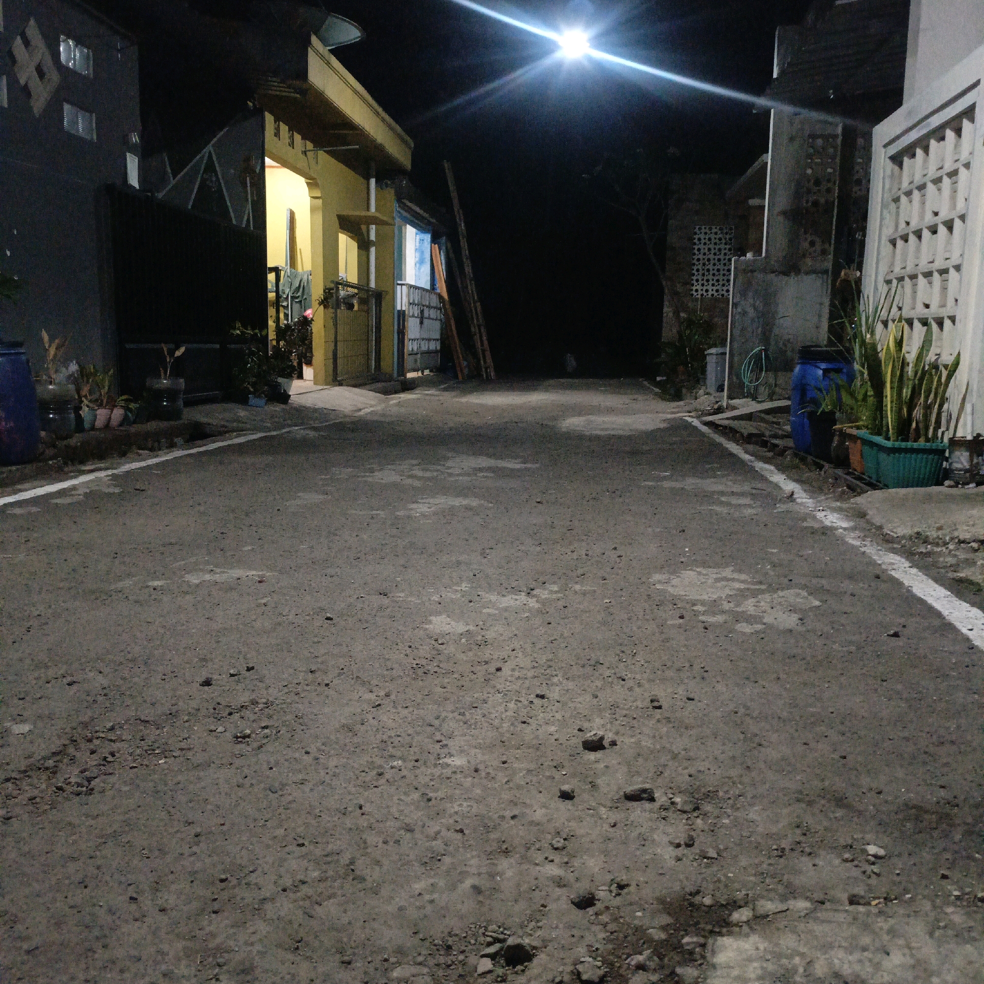 The image depicts a dimly lit outdoor scene, likely a residential street or alleyway at night in Salatiga, Indonesia. The foreground is dominated by a rough asphalt road with scattered pebbles and some lighter patches. A white line marks the edge of the road on the right side.
On the left side of the street, a dark building with visible windows is present, partially obscured by a dark fence or wall. Several potted plants are visible in front of this structure, some in blue barrels and others in smaller pots.
To the right of the street, there is a lighter-colored building with a yellow facade and an overhang. A metal fence runs along the front of this property, and laundry is hanging on a rack behind it. Further down the street, a ladder leans against a wall.
On the far right of the image, a light-colored structure with a decorative, perforated facade is visible. In front of this, a green planter holds several tall, spiky plants, possibly Sansevieria (snake plants). A blue barrel is also situated near these plants.
The overall lighting is dim, with a bright street lamp at the top center of the image casting strong illumination and creating lens flares. The background is dark, suggesting nighttime and minimal ambient light. No people are visible in the image, and no specific actions or interactions are depicted. The scene appears to be a static view of a quiet, possibly residential area in Salatiga.