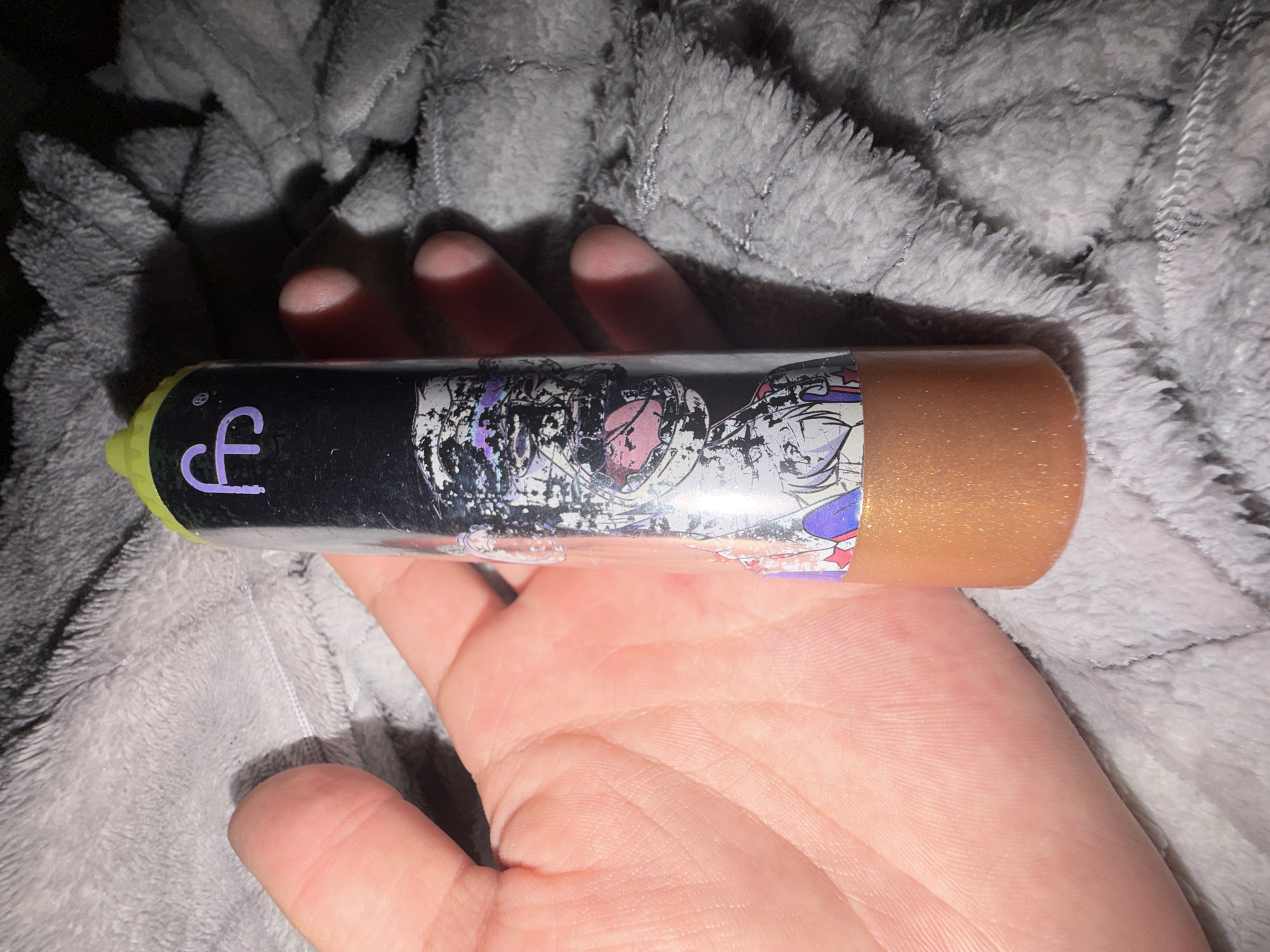 A person's hand is holding a clear cylindrical object with a lime green cap. The object is adorned with a black label featuring a purple 'ff' logo and an anime-style graphic of a character with a pink tongue. The lower portion of the object contains a shimmering golden liquid. The object is placed on a soft, grey, fuzzy textile surface. The image appears to be taken in an indoor setting, with no specific identifiable landmarks from Dorsten, Germany.