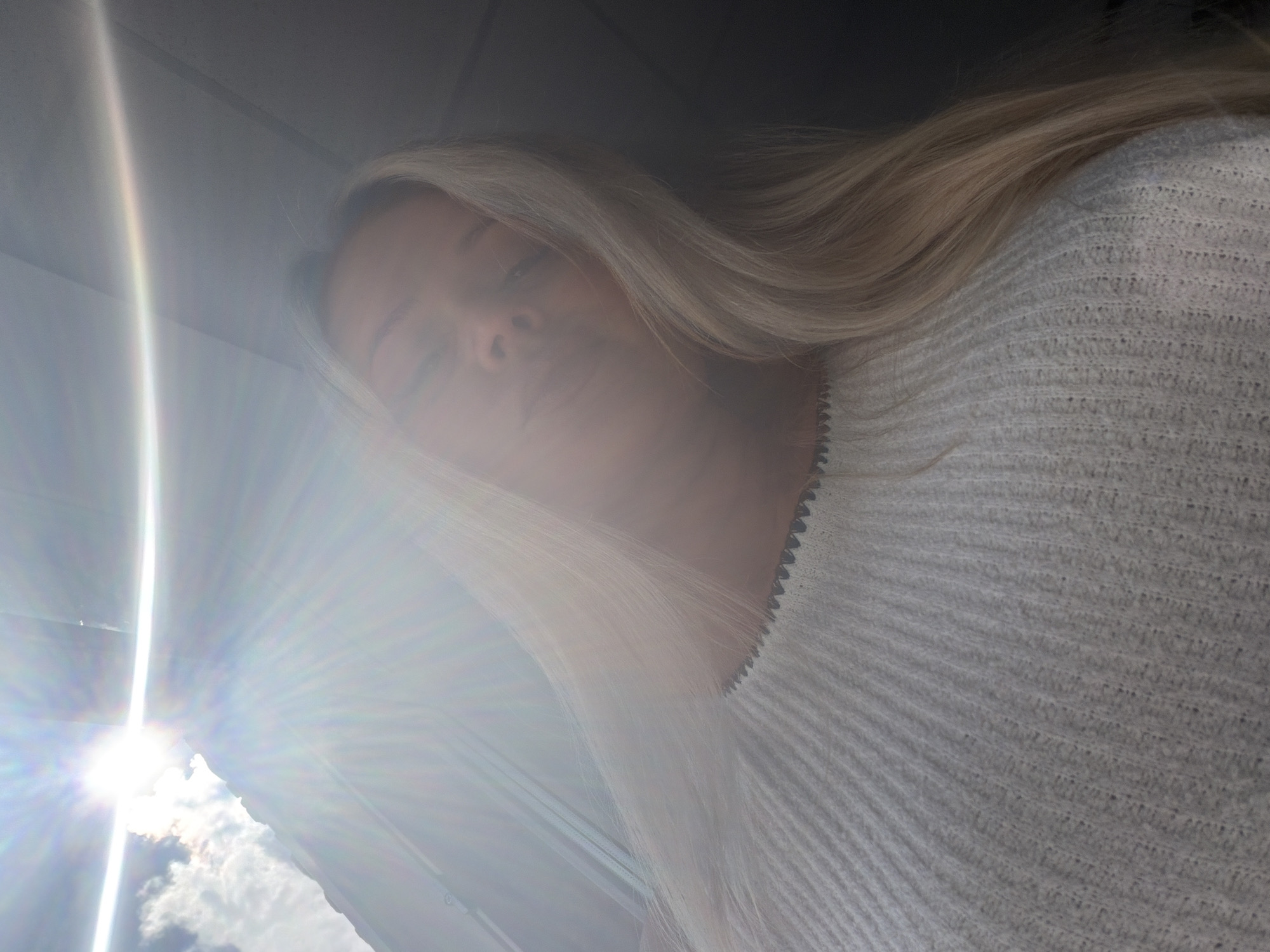 The image depicts a young woman with blonde hair wearing a white ribbed sweater. She is looking upwards and slightly to the side, with her face partially obscured by bright light flares emanating from the sun. The lighting suggests an outdoor setting, possibly on a sunny day, with clouds visible in the upper left corner. The composition is a close-up, focusing on the woman's upper body and face. There is no information in the image to suggest a specific location in Sarapul, Russia.
