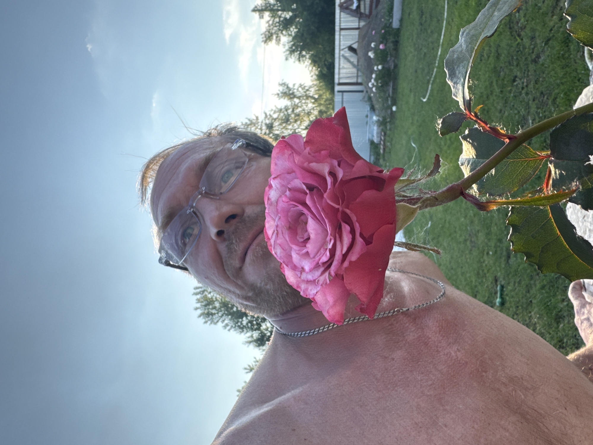 A close-up, low-angle, outdoor selfie shows a man in his 50s or 60s holding a pink rose up to his face. He is wearing glasses and a silver chain necklace. The man's upper body and shoulder are visible, and he has light skin with some redness, possibly from sun exposure. The background shows a grassy yard with a fence, trees, and a clear blue sky with a few wispy clouds. The scene appears to be set in a rural or suburban area, possibly in the city of Chaikovsky, Russia, given the context of the request. The lighting suggests it is late afternoon or early evening.