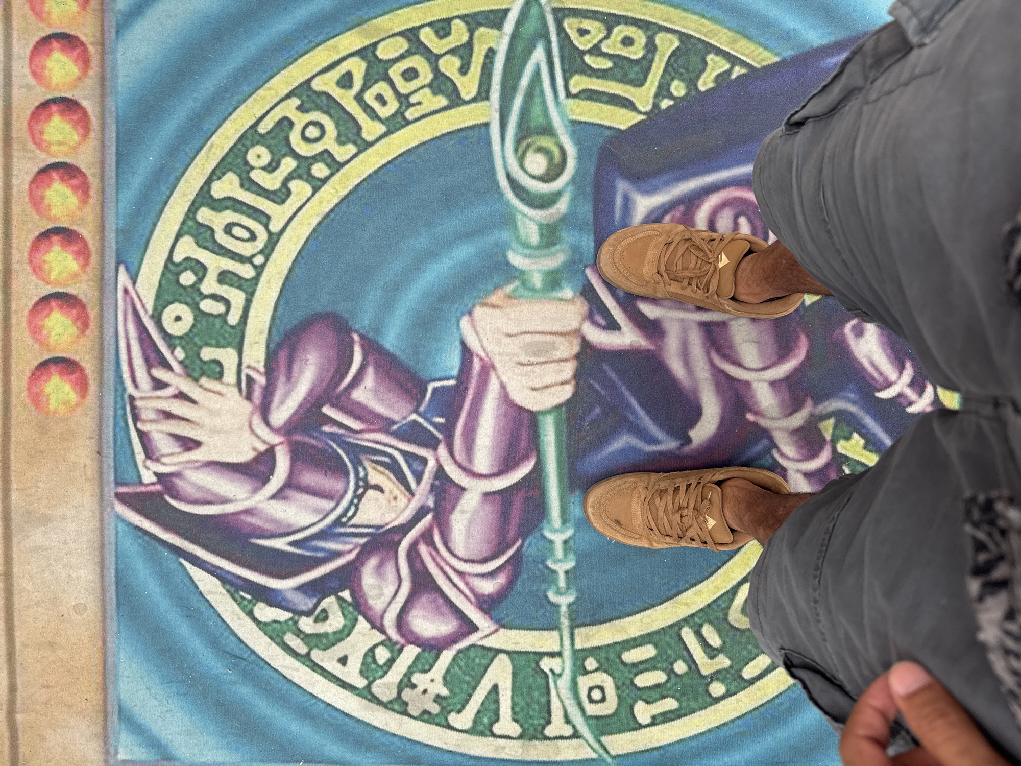 The image shows a person's legs and feet standing on a large mat featuring the Yu-Gi-Oh! card "Dark Magician." The mat is blue with a circular green and yellow design, and the Dark Magician character is depicted in purple and blue robes, holding a staff. The person is wearing grey cargo shorts and tan sneakers. The background of the image shows the top border of the mat, which has a series of small red circles with yellow arrows. The scene appears to be indoors, on a flat surface. There is no discernible El Paso, United States of America specific location information in the image.