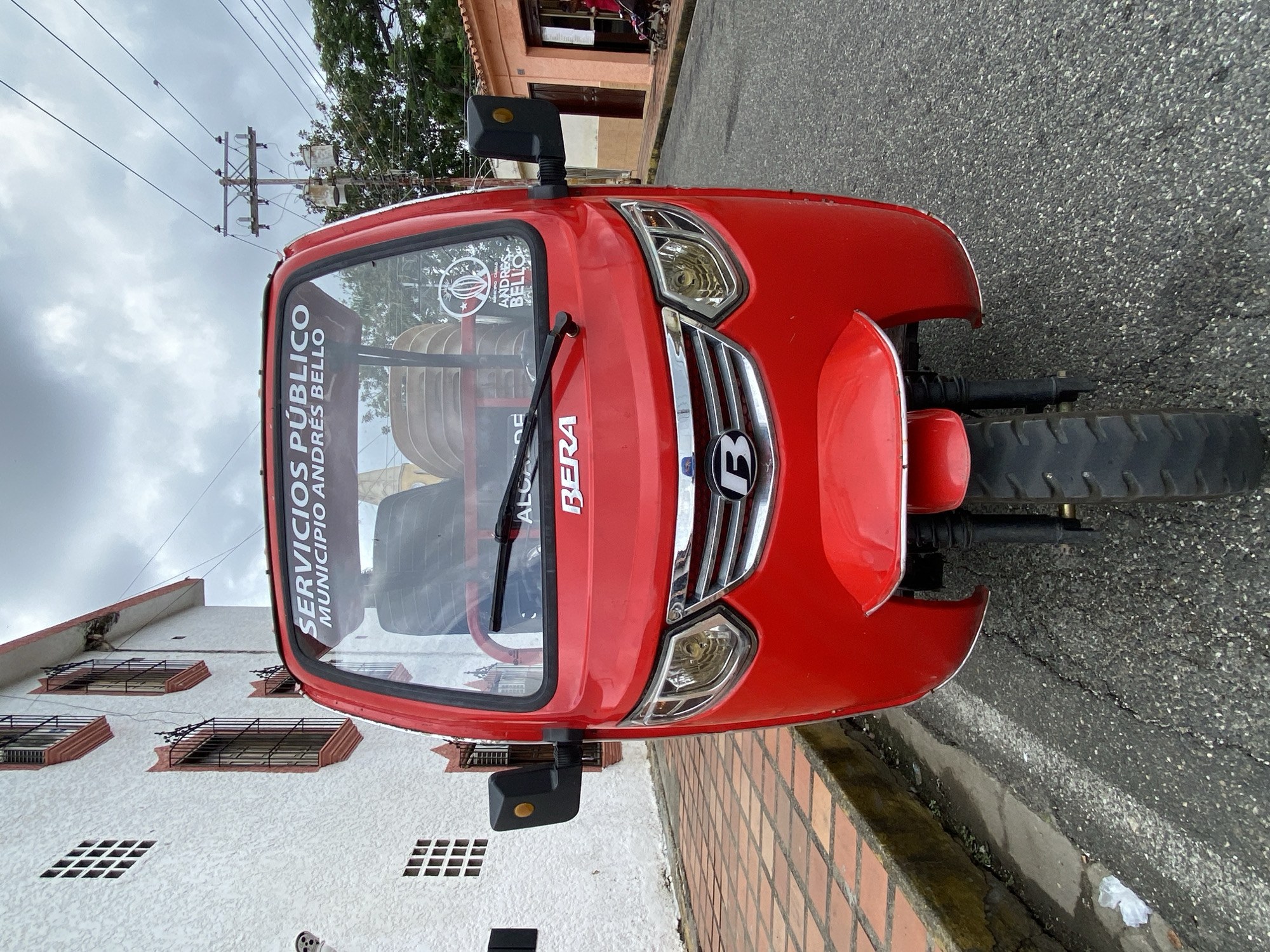 The image depicts a red three-wheeled motorized vehicle, a mototaxi or motocarro, centrally positioned on an asphalt street, facing the viewer. The vehicle's windshield displays white lettering: "SERVICIOS PÚBLICOS MUNICIPIO ANDRÉS BELLO" and "ALCALDÍA." The brand "BERA" is visible in red on the front cowl. A "B" logo is on the chrome grille. Stacked white plastic chairs are visible through the windshield.
To the left, a multi-story building with textured white stucco walls features multiple windows with decorative reddish-orange trim and black iron grates. A section of a tiled sidewalk, composed of reddish-brown square pavers, runs along this building. A white, skull-like decoration is partially visible on the building's lower left side.
To the right of the vehicle, the asphalt street continues. In the mid-ground, a building with a peach-colored facade is visible. A person in red attire is seated in a stroller near the entrance of this building. Overhead, against an overcast sky, utility poles with multiple wires traverse the scene. Green tree foliage is visible in the background. The scene is located in the city of San José de Barlovento, within the Andrés Bello Municipality, Venezuela.