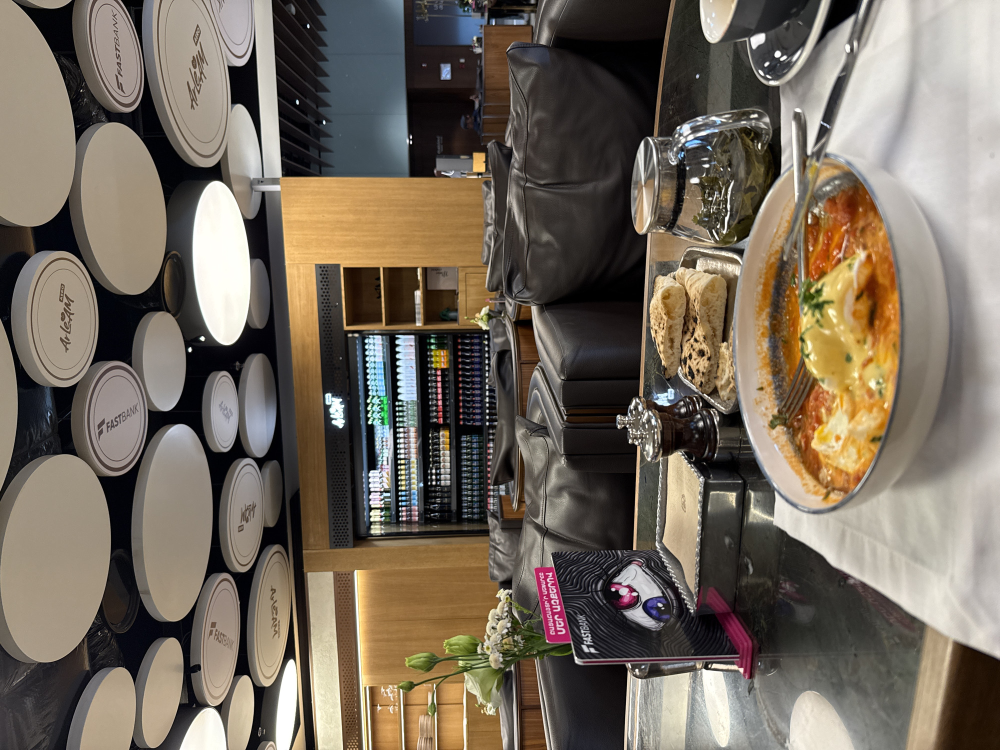 The image depicts an interior scene, likely a lounge or waiting area, featuring a table set with food and drinks. In the foreground, a white bowl contains a dish of eggs in tomato sauce, garnished with herbs. To the right of the bowl is a black teacup and saucer. Behind the bowl, a metal tray holds several pieces of flatbread. A glass teapot filled with green tea sits to the right of the tray.
A sign with the "FASTBANK" logo and an abstract eye design is prominently displayed on the table. A small bouquet of white flowers with green buds is placed to the left of the sign. In the background, dark leather armchairs are arranged. Beyond the seating area, a refrigerated display case filled with bottles of beverages is visible, along with wooden shelving and cabinetry. The ceiling is decorated with large, circular, disc-like fixtures. One of these discs bears the "FASTBANK" logo, while others have text that appears to be "ArLeAm" and "ArLeAm AZBO". This suggests the location is associated with Fast Bank, a financial institution in Yerevan, Armenia, and possibly a lounge area branded with these entities. The overall impression is one of a comfortable and well-appointed public space.