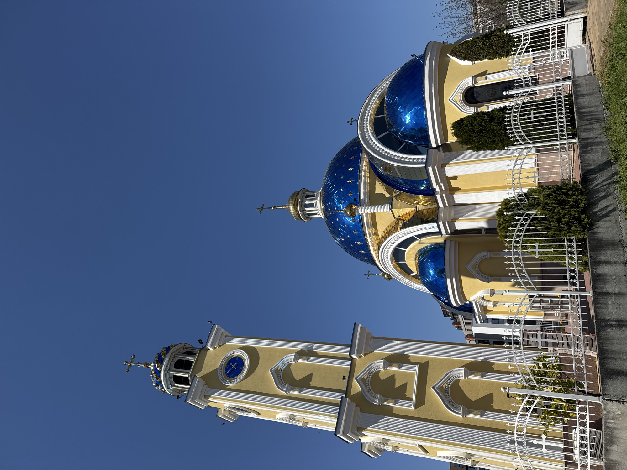 The image shows the Dormition of the Mother of God Cathedral in Plovdiv, Bulgaria. It's a low-angle shot capturing the church's architectural elements against a clear blue sky. The cathedral features a tall bell tower with a clock and distinctive yellow and white detailing. The main structure of the church displays several blue domes with gold accents. A white wrought-iron fence surrounds part of the cathedral grounds, with some greenery present. The scene appears to be taken during the day, likely under sunny conditions. There is no visible sign of people.