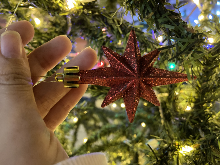 A hand delicately holds a sparkling, red star ornament. The ornament, with its eight points and glittery surface, features a gold-colored cap. The backdrop is a Christmas tree illuminated with twinkling lights, both white and colored. Green needles of the tree and the soft focus of the lights create a festive and warm atmosphere.