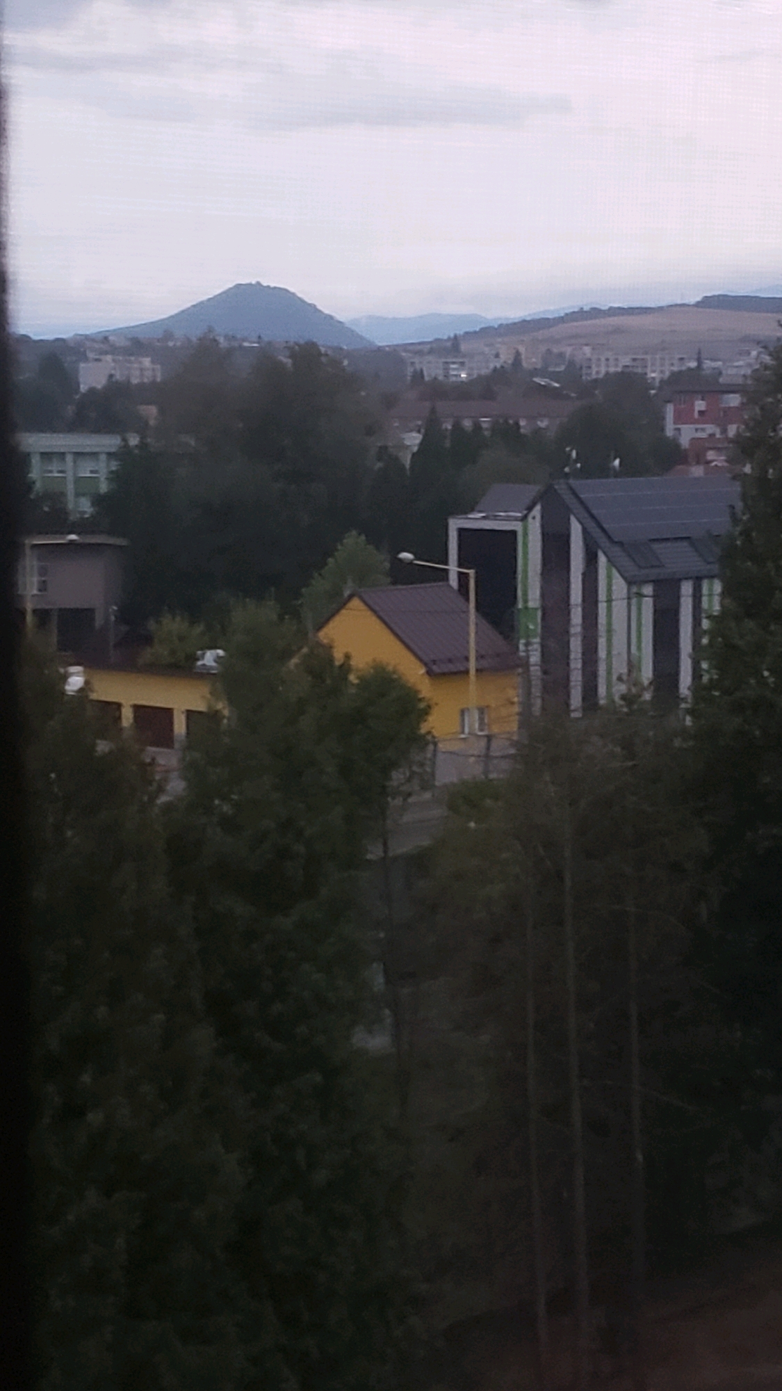 The image is an outdoor, slightly hazy, daylight view of a residential area in Prešov, Slovakia, with a prominent hill in the background. In the foreground, several buildings are visible, including a bright yellow house with a dark roof and two windows. Adjacent to it is a modern building with a black and white striped facade. Trees and foliage obscure parts of the scene. In the middle ground, there are other residential buildings, some with darker roofs and lighter walls. The background features a distinctive, conical hill with what appears to be a structure on its summit, and rolling hills beyond it. The sky is overcast with diffused light.