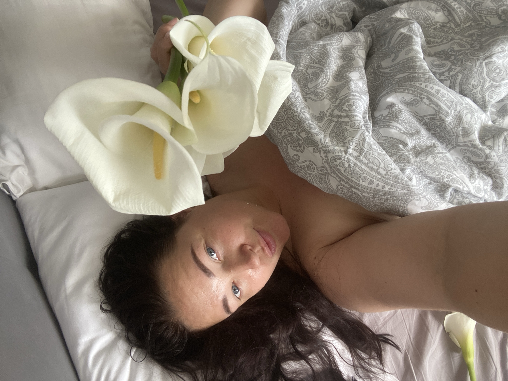 A woman with dark hair and blue eyes lies in bed, holding a bouquet of white calla lilies. She is wearing a grey and white paisley patterned duvet. The room appears to be a bedroom in Sochi, Russia, with soft lighting.